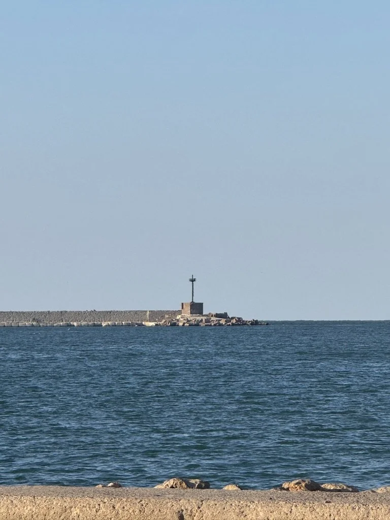 A calm ocean with a breakwater and a lighthouse on a small pier in the distance, under a clear blue sky.