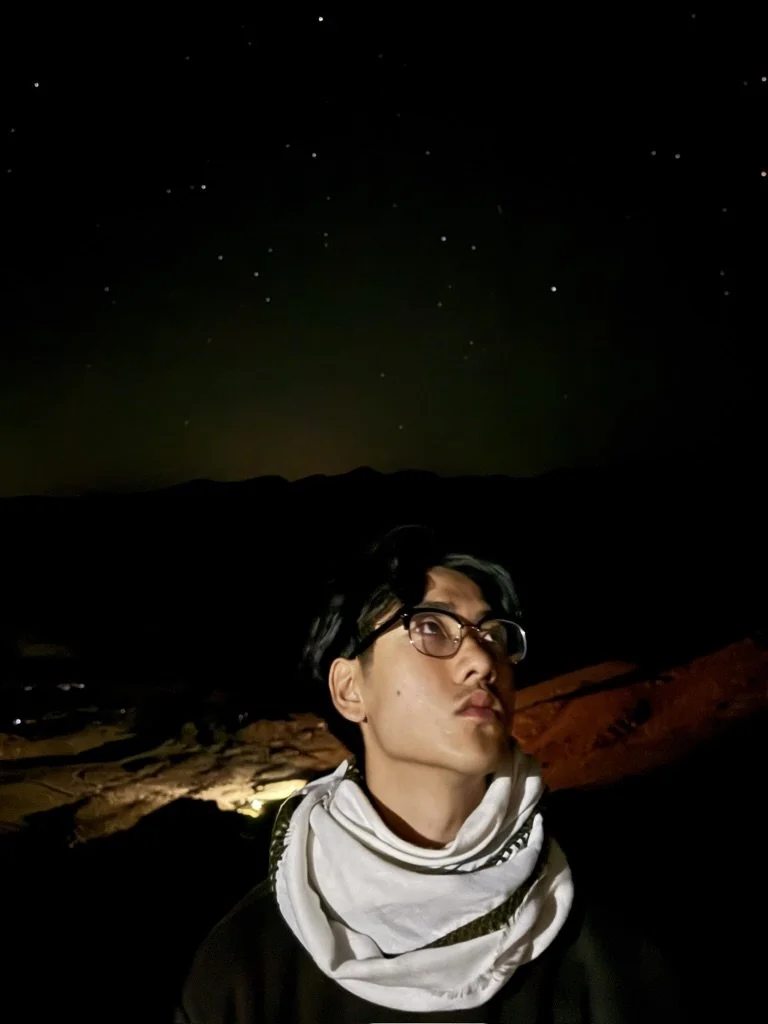 A person with short black hair and glasses looking upwards, wearing a white scarf around their neck, standing in a dark outdoor setting at night with a starry sky and silhouette of mountains in the background.