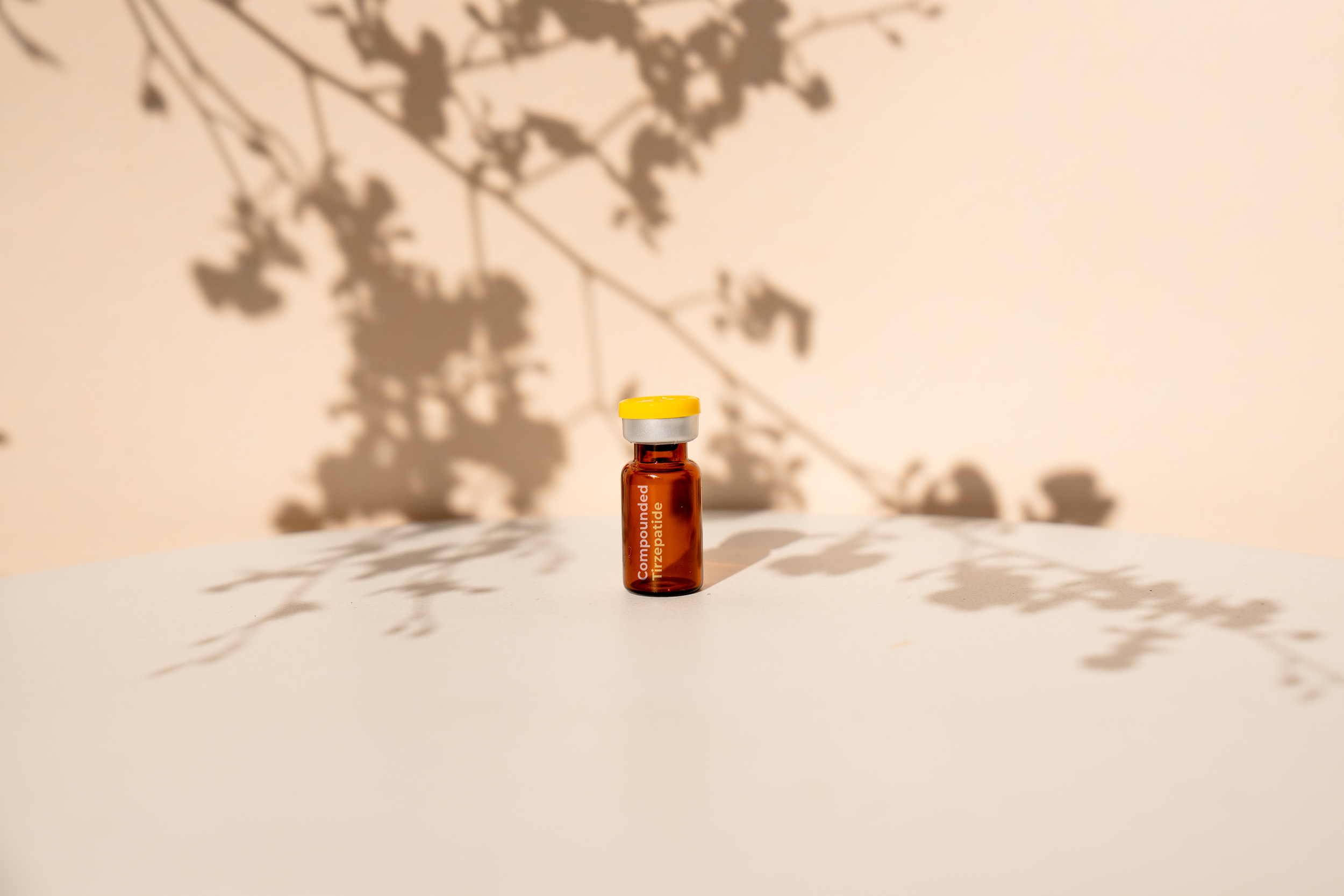 A small amber glass vial with a yellow cap, labeled 'Compounded Titration' is placed on a white surface. Shadows of leaves are cast on the wall behind it, creating a natural pattern.