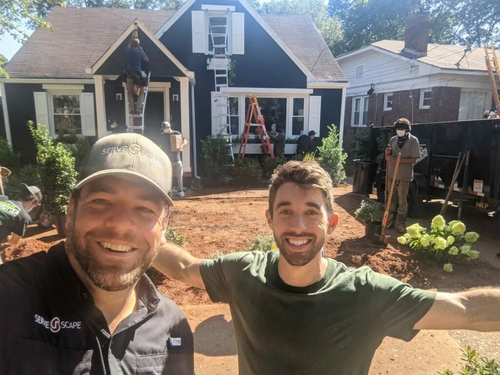 Two smiling men take a selfie in front of a house under renovation, with workers planting bushes, digging, and working on the house outside.