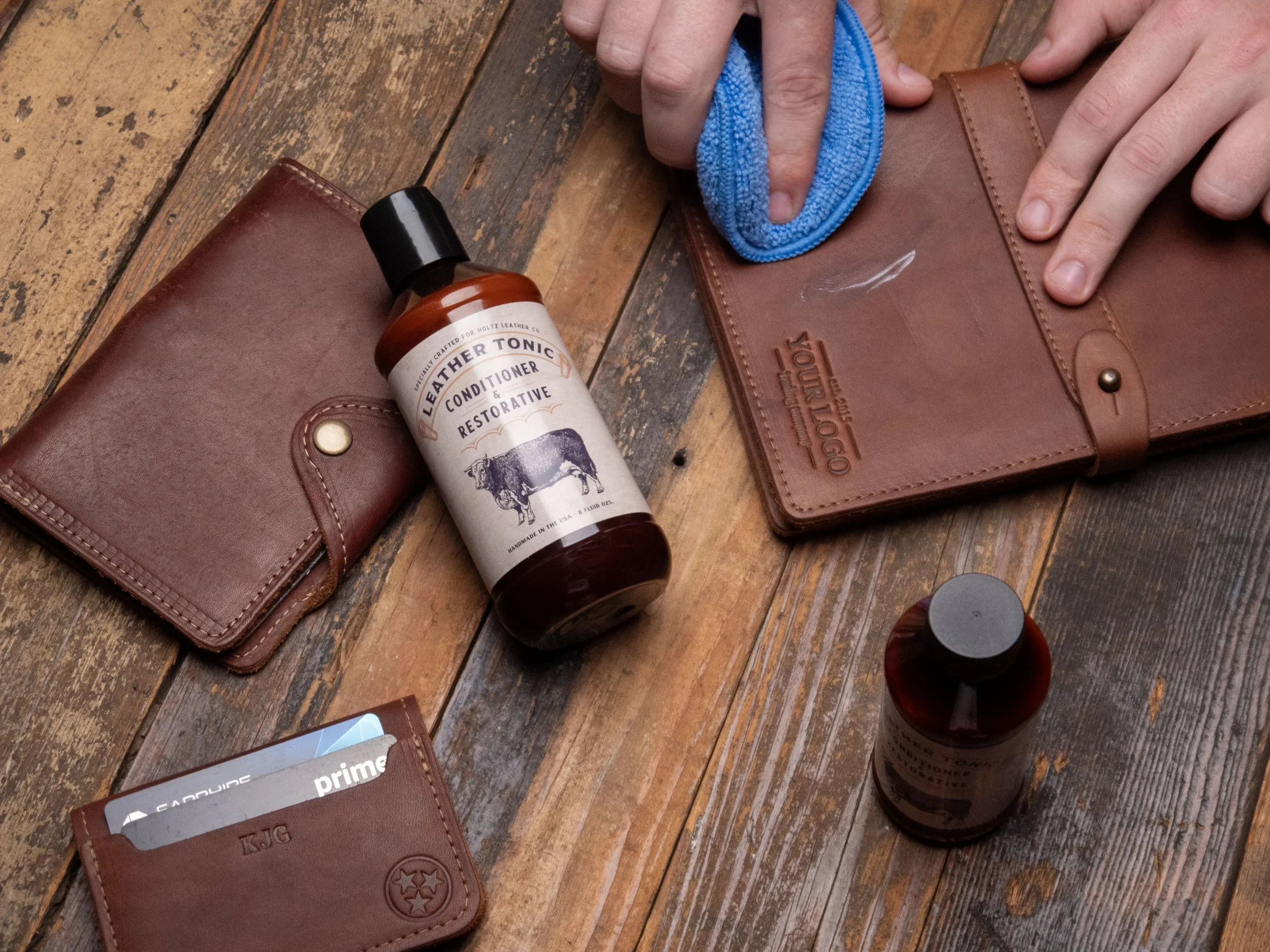 Cleaning and conditioning leather goods, such as wallets and folders, on a wooden table with a blue cloth and leather conditioner bottles.