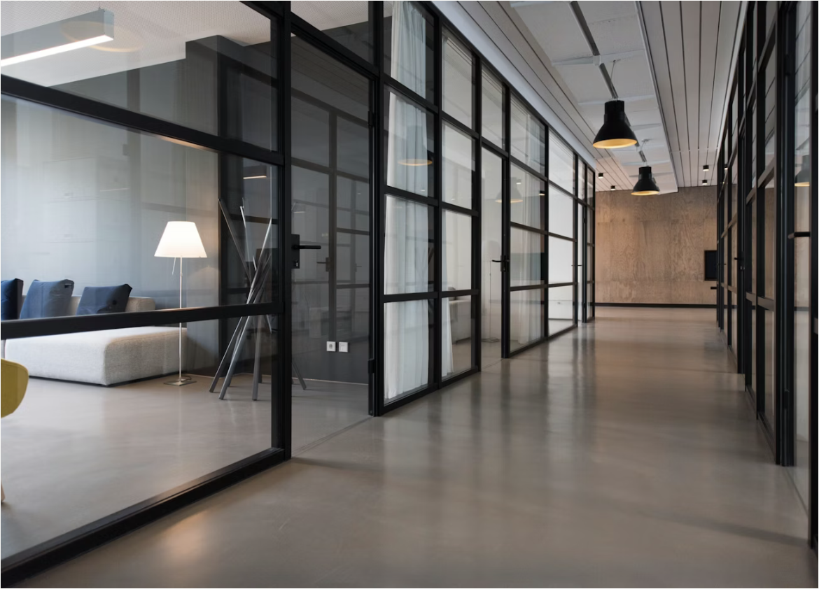 Modern office corridor with glass walls, black frames, and black hanging lights. A seating area with a white sofa, blue pillows, a floor lamp, and a small table are visible through the glass walls.