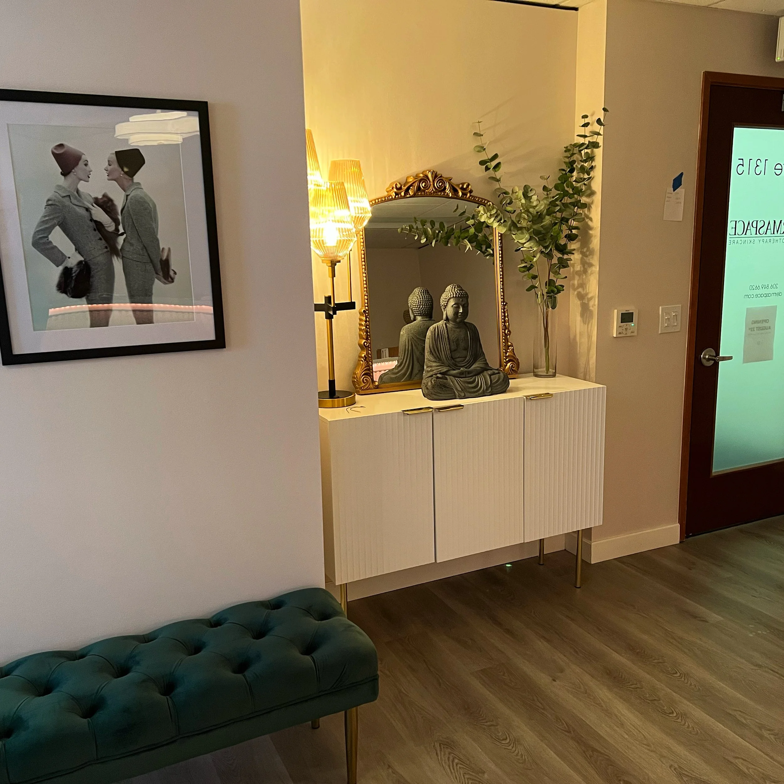 A corner of a room decorated with a large framed black-and-white photograph of two women facing each other, a tall wooden floor lamp with a warm glow, a gilded mirror, a small Buddha statue, and a glass vase with eucalyptus branches. There is also a partially visible green tufted ottoman and a doorway with a glass door displaying green text.