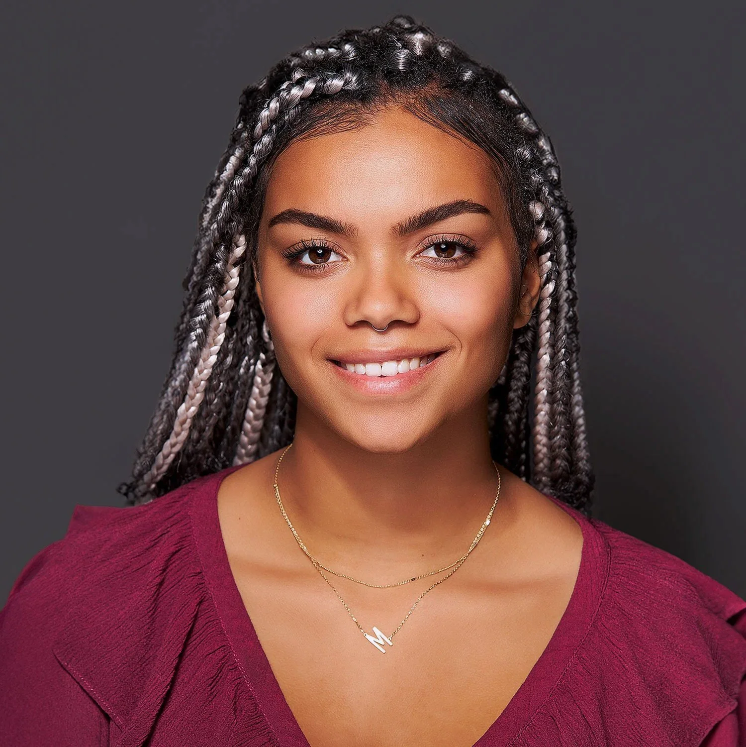 A woman with braided hair, smiling, wearing a maroon top and layered necklaces, against a dark gray background.