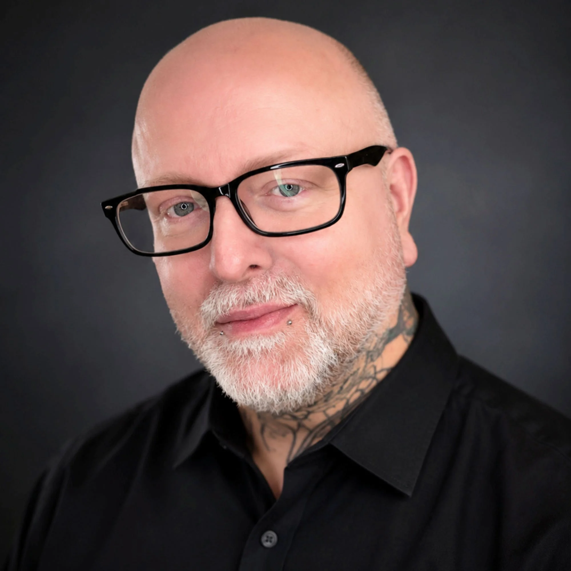 Close-up of a man with light skin, a beard, and a tattoo on his neck, wearing black-rimmed glasses and a black shirt, against a dark background.