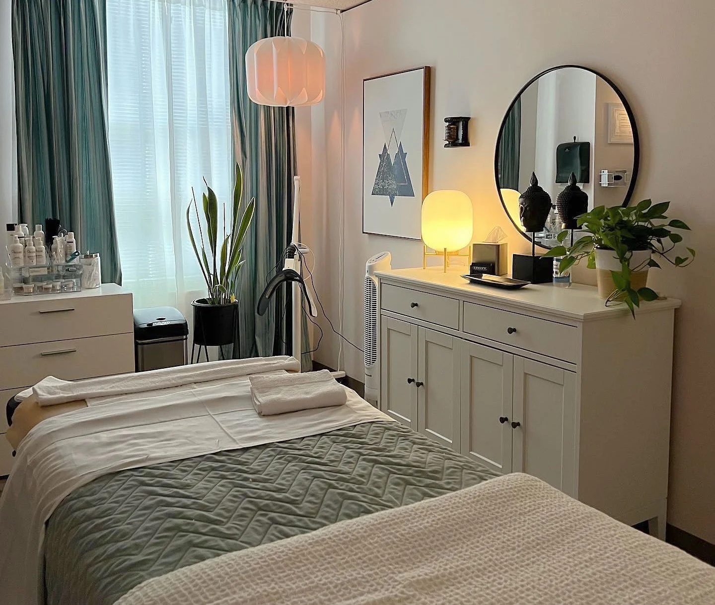 A cozy bedroom with a bed, a large mirror, a dresser with decorative objects and a lamp, a tall potted plant, and a window with teal curtains.