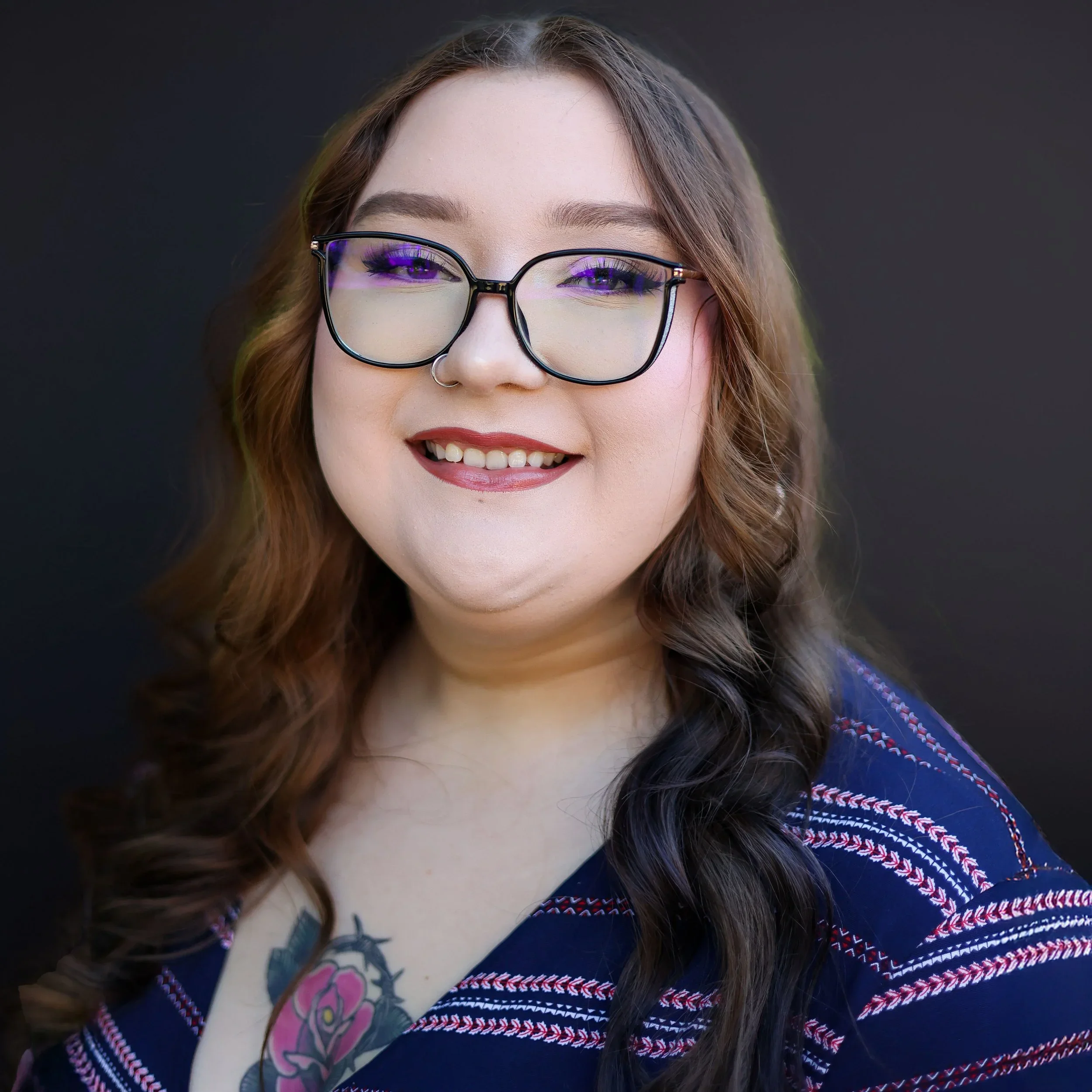 Close-up of a smiling woman with long brown hair, wearing glasses, a nose ring, and a multi-colored, patterned top, against a dark background.