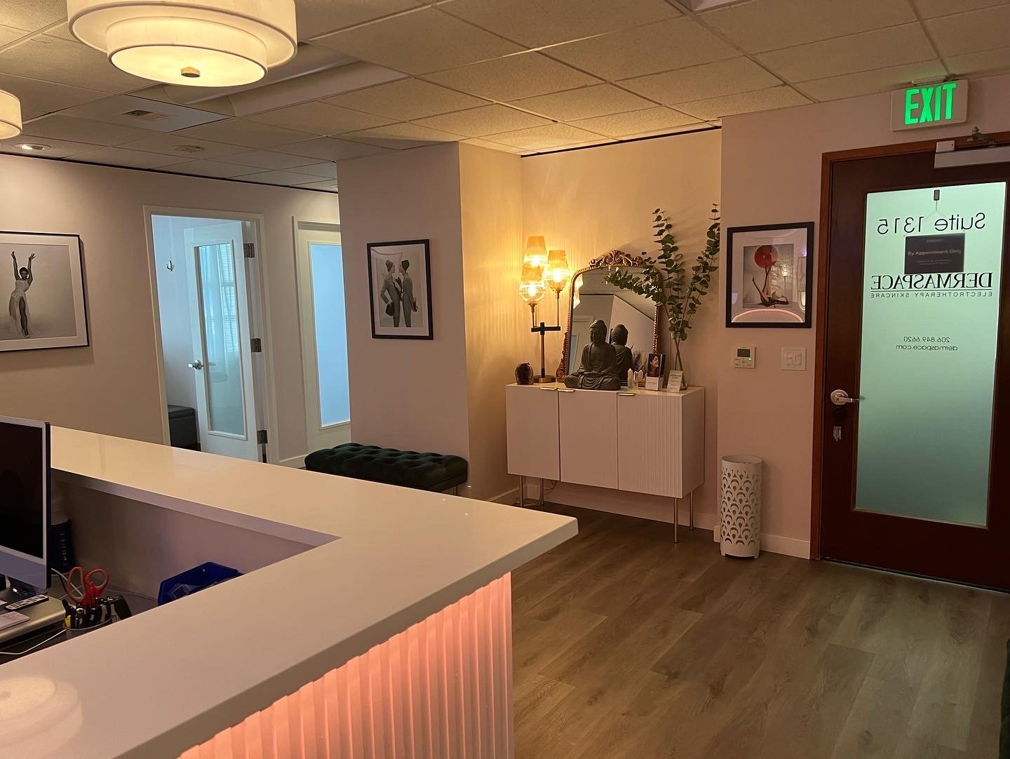 Reception area with a white counter, framed art on the walls, a white console table with a Buddha statue, plants, and framed photos, a mirror, a modern light fixture, and glass doors leading to rooms, with furniture and decorations creating a calming atmosphere.