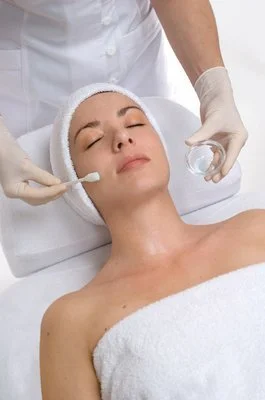 A woman receiving a facial treatment at a spa or clinic, lying on a treatment bed with closed eyes, wearing a white headband, while a professional applies a skincare product with a brush.