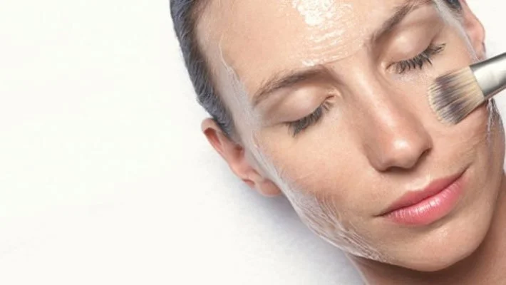 A woman with her eyes closed applying a facial mask with a brush.