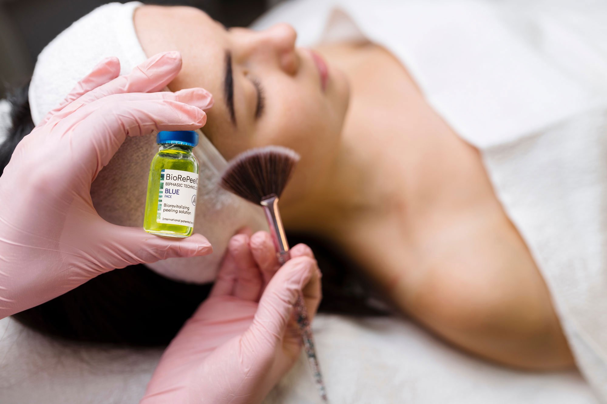 A woman receiving a facial treatment while lying down with her eyes closed, wearing a headband. A practitioner wearing pink gloves holds a small glass bottle labeled 'BioRePeel' and a brush, applying a treatment on the woman's face.