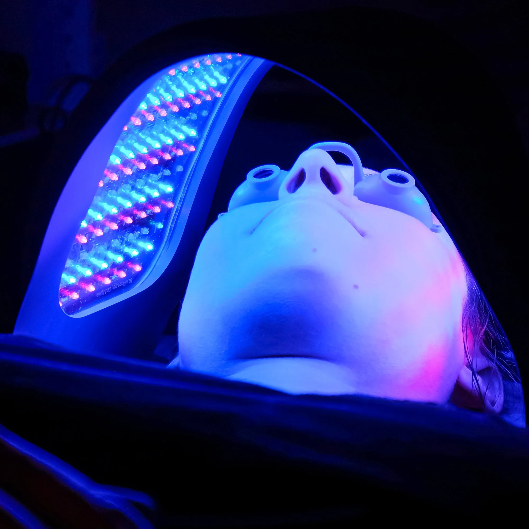 A person lying on a medical scanner table underneath a circular MRI machine with bright blue, pink, and purple lights illuminating the scene.