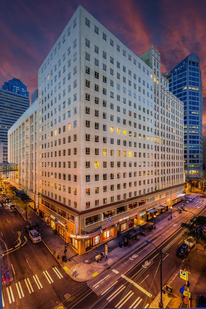 Tall city building at dusk with illuminated windows and traffic moving on the street below.