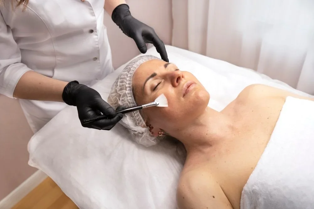 A woman receives a facial treatment in a spa or clinic setting, lying on a bed with her eyes closed, wearing a hair cap, while a skincare professional applies a product with a brush.