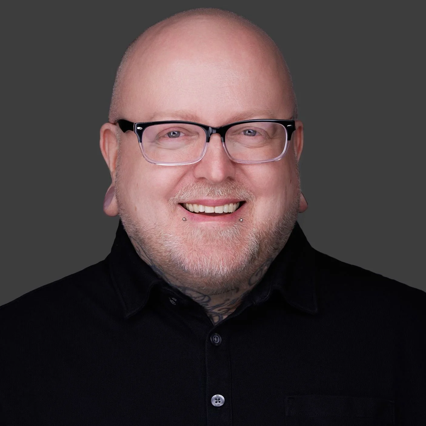 Portrait of a smiling bald man with glasses, facial piercings, and tattoos, wearing a black shirt against a dark gray background.