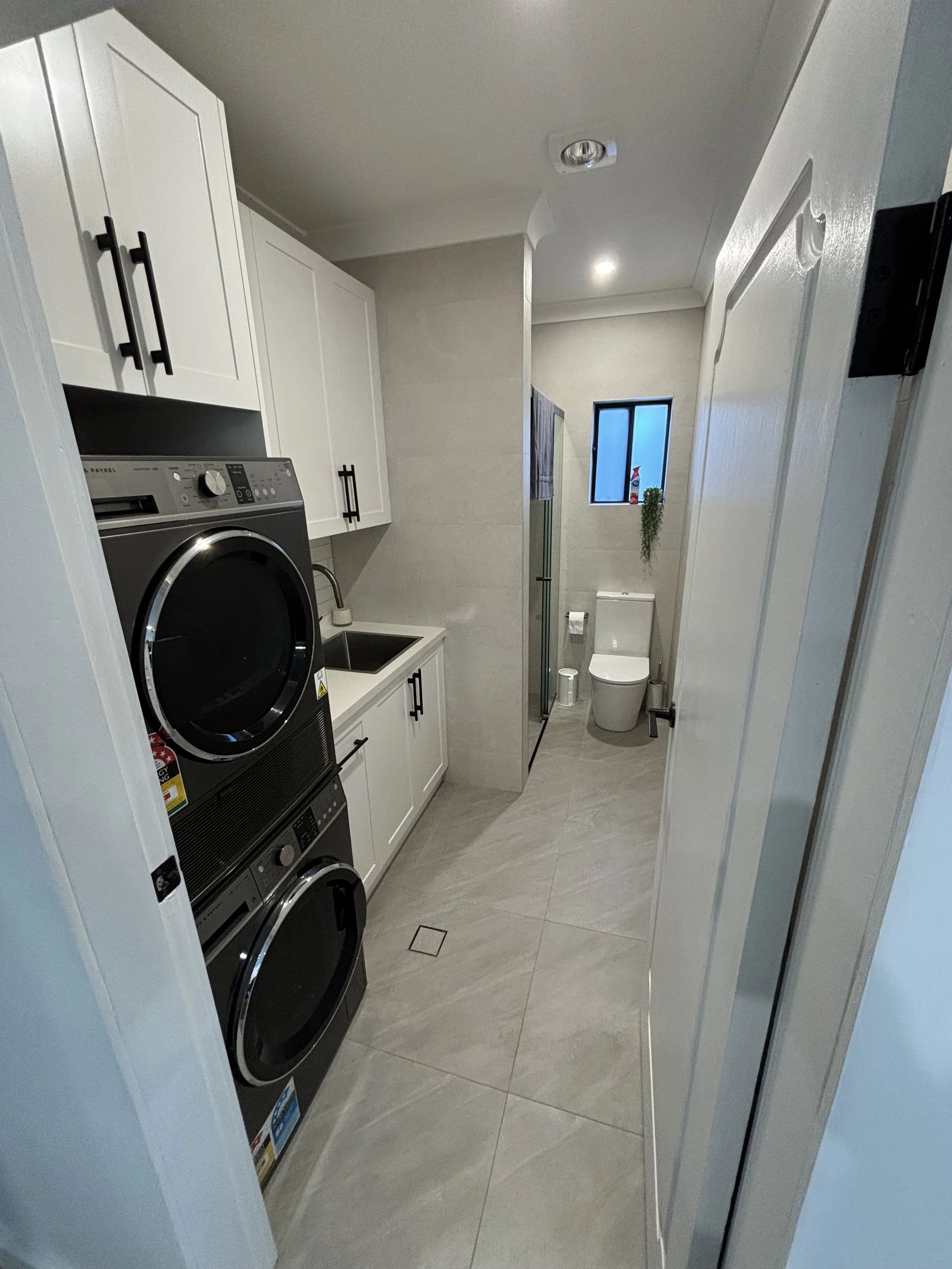 A compact laundry room with white cabinets, a washing machine and dryer, a small sink, and a toilet. There is a window with a small plant and a spray bottle, beige tiled floor, and light-colored walls.
