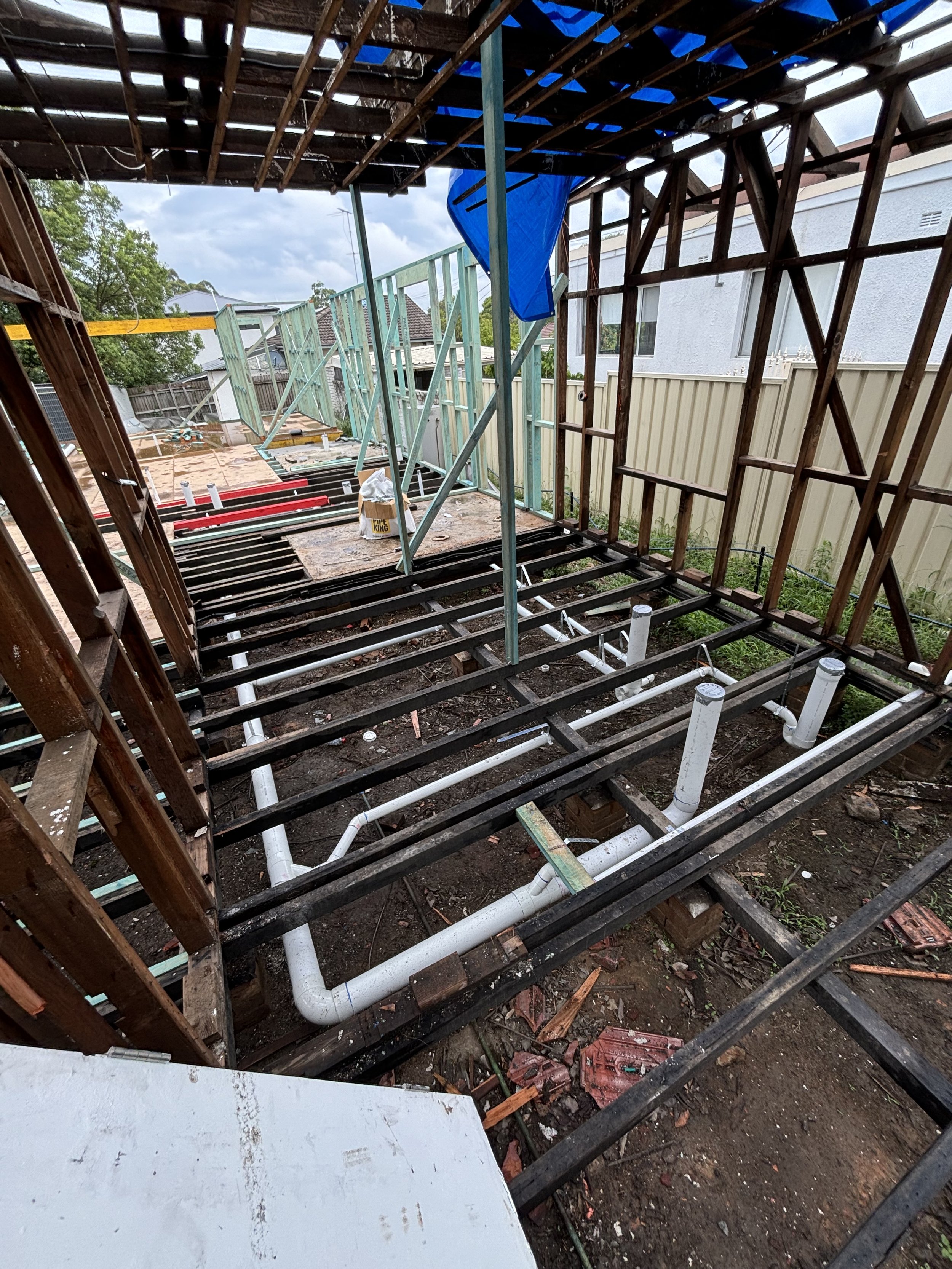 A house under construction with exposed wooden framing and plumbing pipes visible on the ground. A blue tarp hangs from the wooden frame, and there is a partially built wall with light green framing. The sky is cloudy, and neighboring houses are visible in the background.