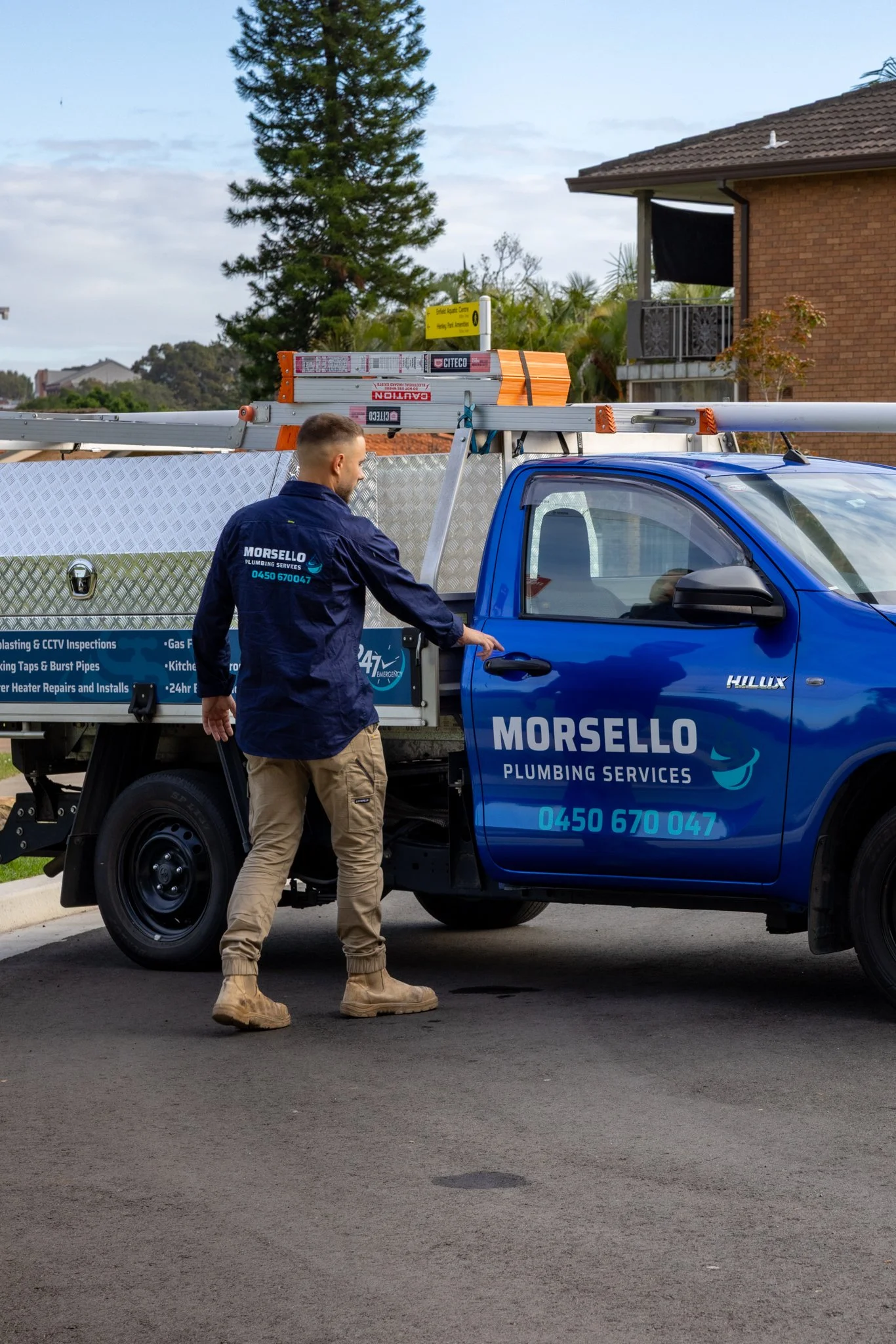 A man in a navy blue shirt and tan pants standing next to a blue plumbing service truck. The man has short hair and is looking down, near the truck's side. The truck is equipped with ladders and tools on its roof, with a yellow sign in the background indicating a nearby aquatic center.