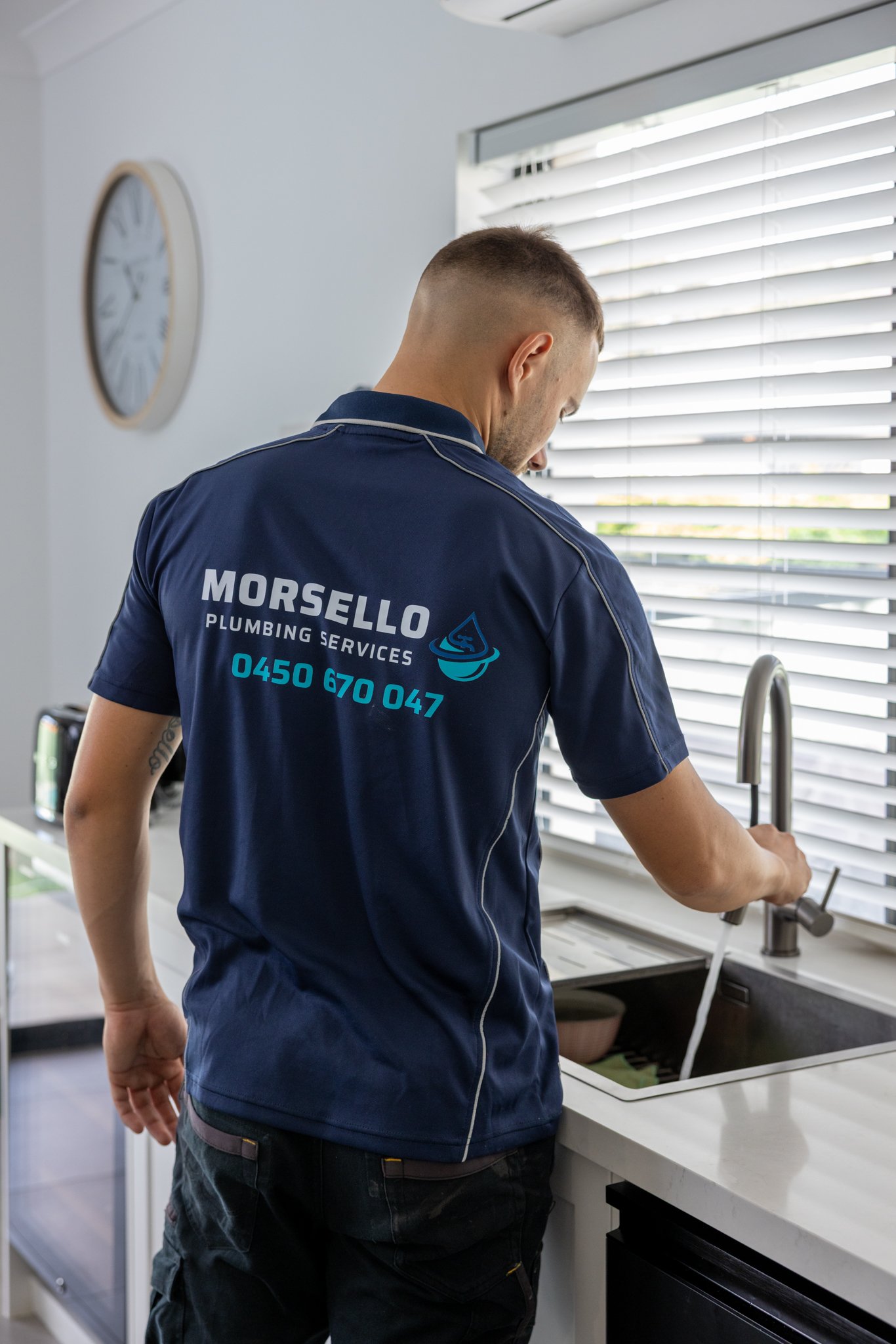 A man with a buzz cut wearing a navy blue shirt with plumbing service branding is at a kitchen sink, working with a faucet under a window with blinds.