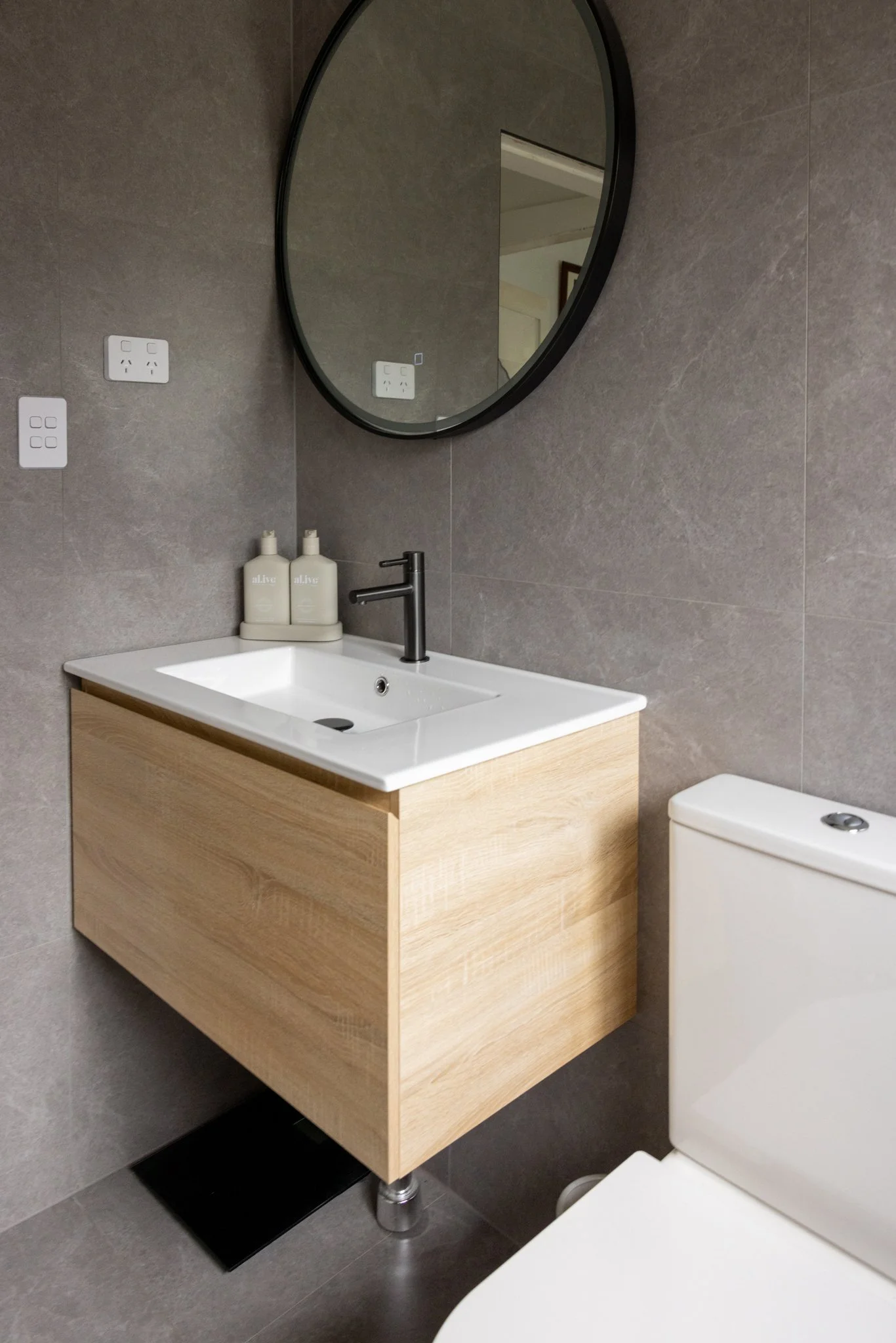 A bathroom with gray tiled walls, a circular mirror, a white sink with a wooden cabinet, two bottles of lotion, a black tap, electrical outlets, and a toilet.