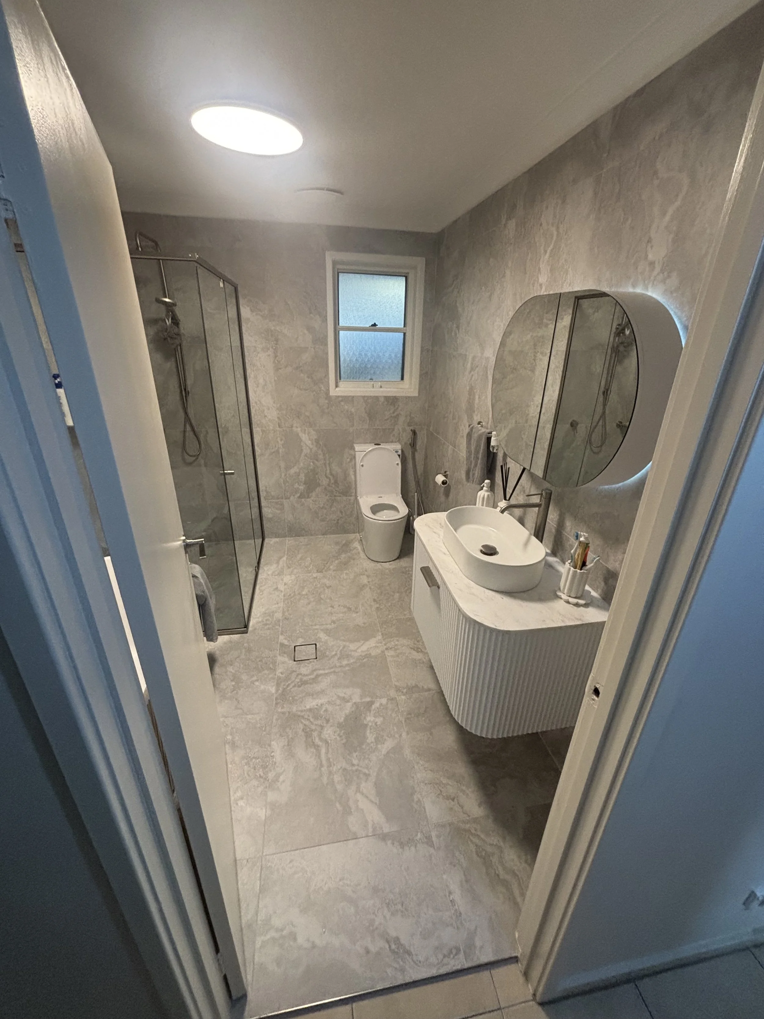 Modern bathroom with gray marble walls and floor, a white toilet, a sink with a curved vanity, a large round mirror, a walk-in shower with a glass door, small window, and ceiling light. Toiletries on a container near sink.