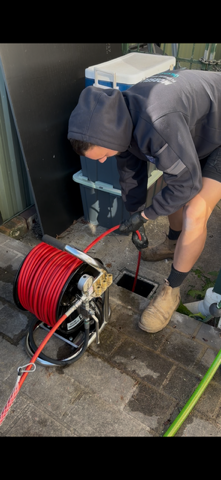 Person in dark clothing and beanie working on outdoor equipment with a red extension cord and hose near a building.