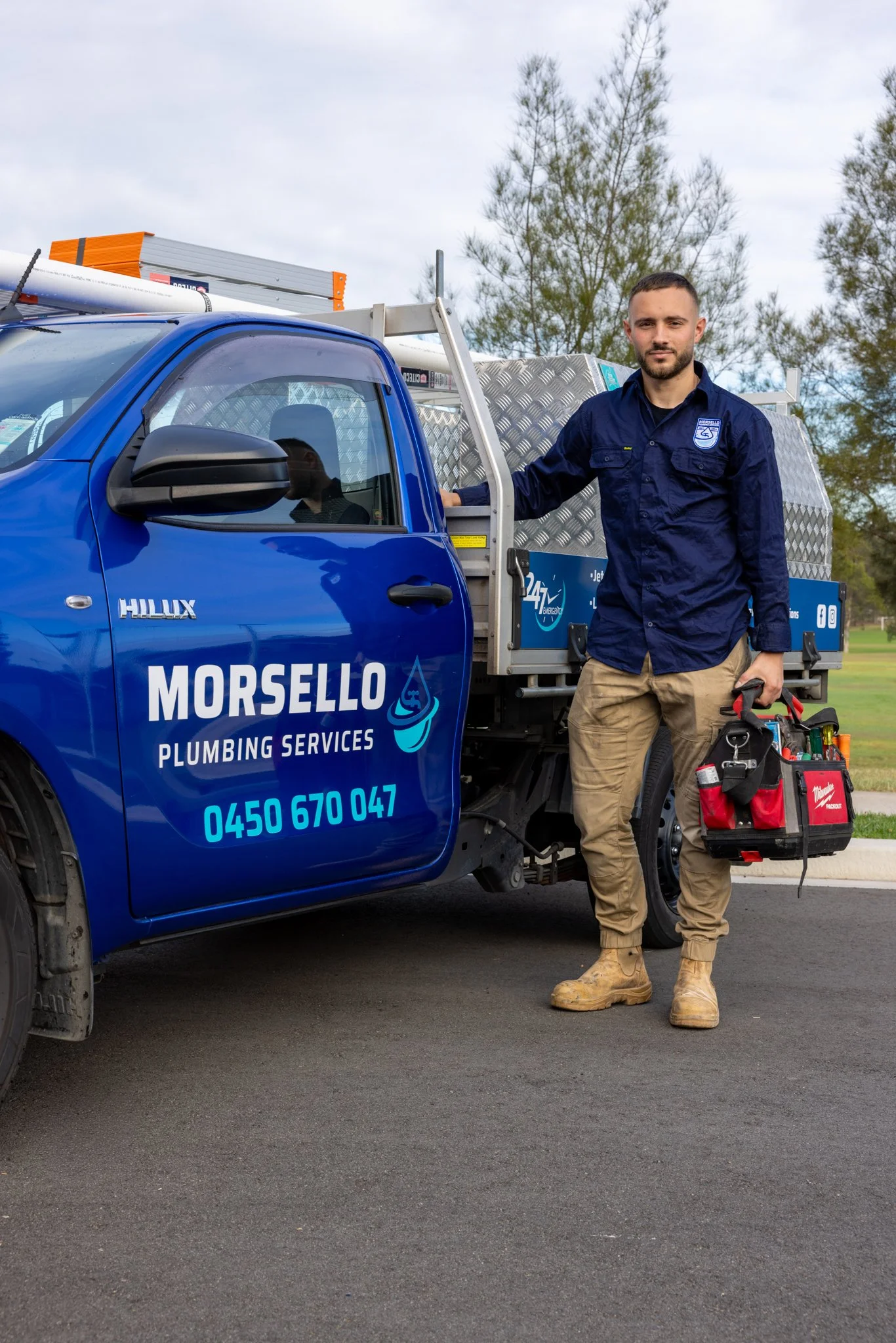A plumber in uniform standing next to a blue Morsello Plumbing Services truck, holding a tool bag with various tools, outdoors on a cloudy day with trees in the background.