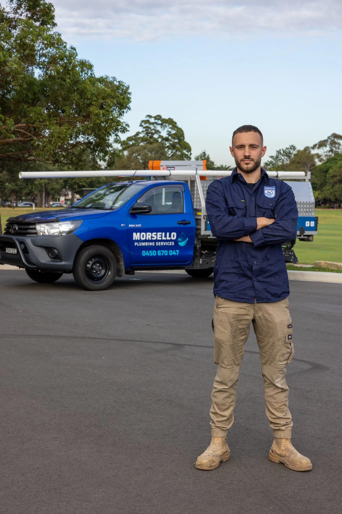 A man standing with arms crossed in front of a blue plumbing service truck with ladders on top, in an outdoor park setting with trees and a cloudy sky.