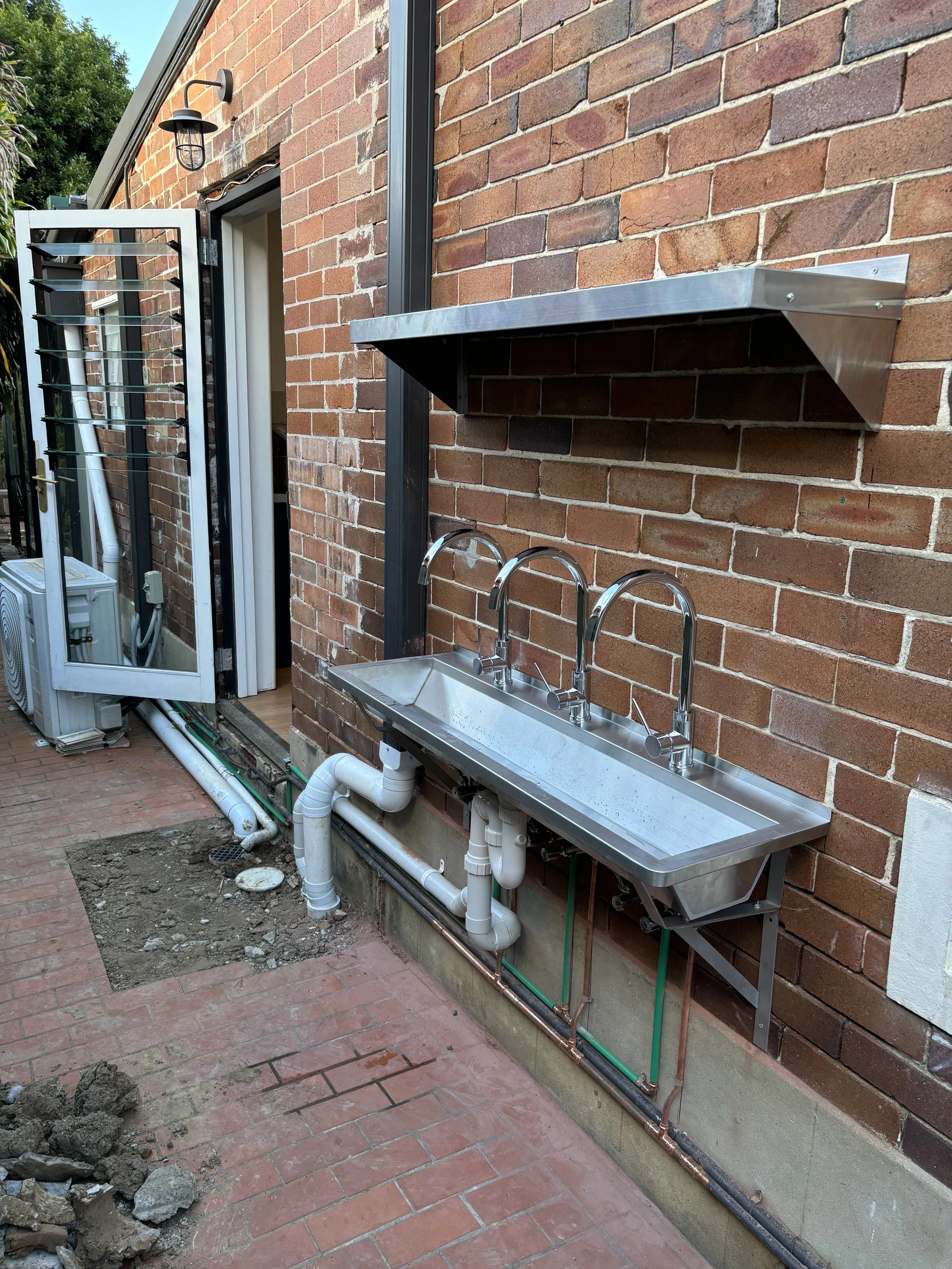 Outdoor brick wall with stainless steel utility sink, a shelf above it, and plumbing pipes beneath. There's a partially open door and a light fixture on the wall. Construction debris and dirt are on the ground.