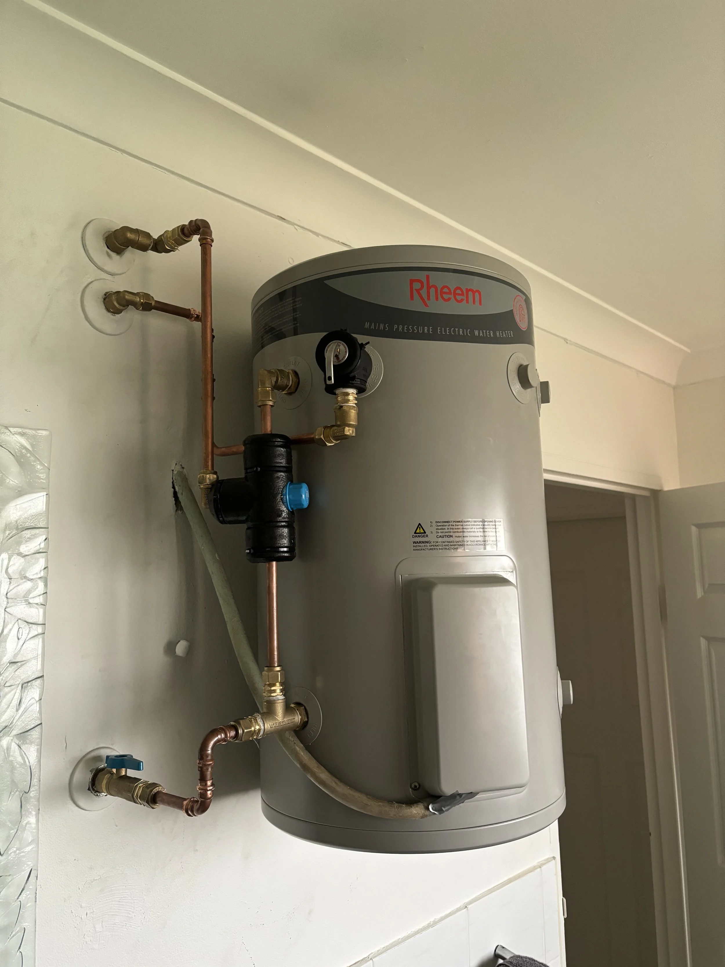 A Rheem electric water heater mounted on a wall with visible copper and PVC plumbing pipes connected to it.