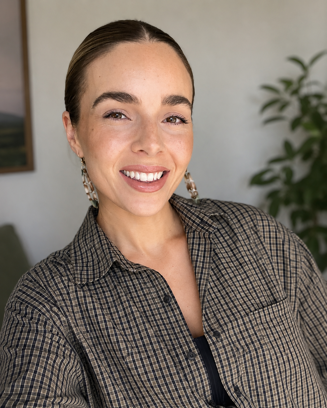 A woman with brown hair slicked back, wearing a plaid shirt and statement earrings, smiling indoors with a plant and a framed picture in the background.