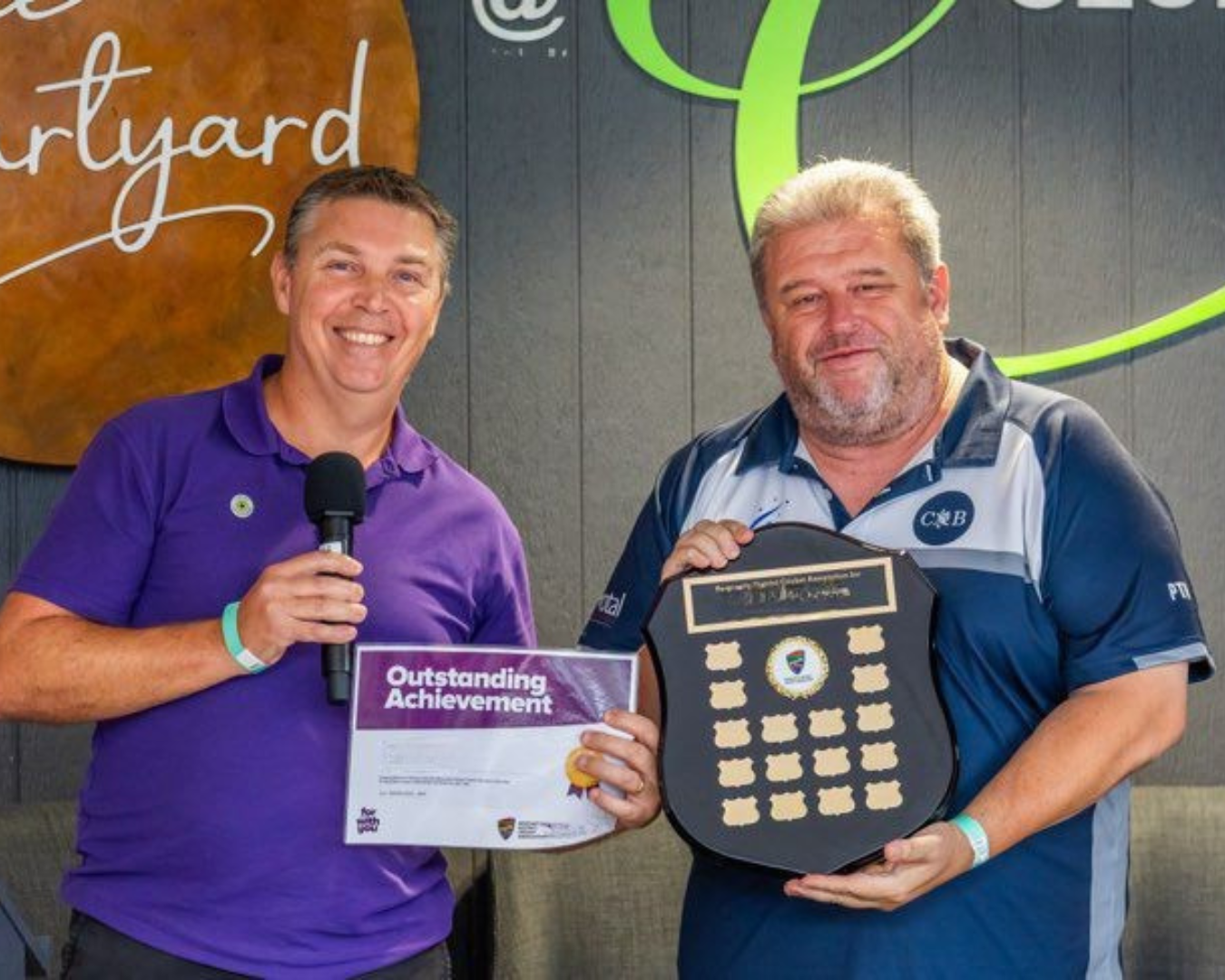 Two men smiling, holding up an award at a community sports event.