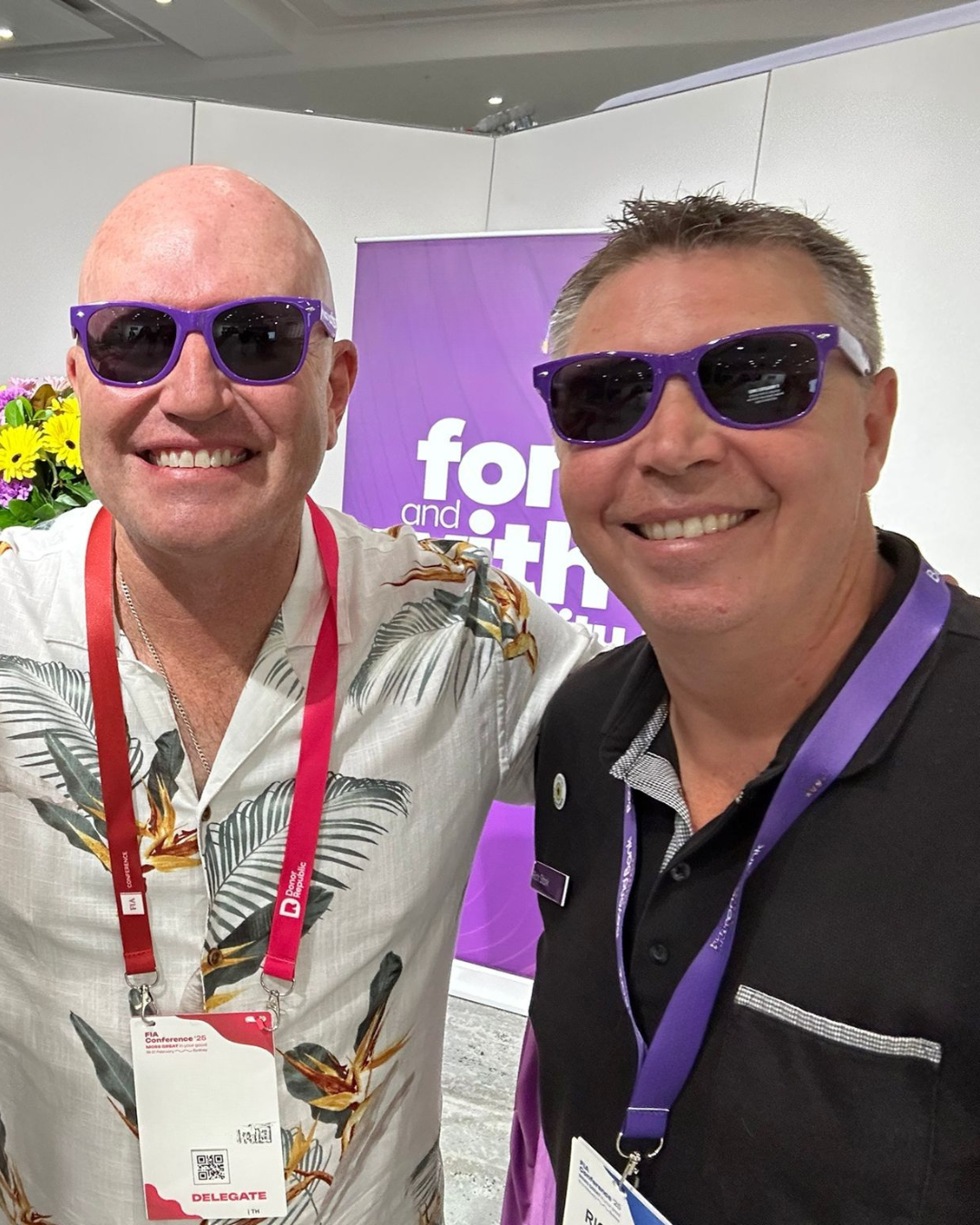 Two men wearing purple sunglasses, smiling, at a fundraising event or conference with a purple backdrop.
