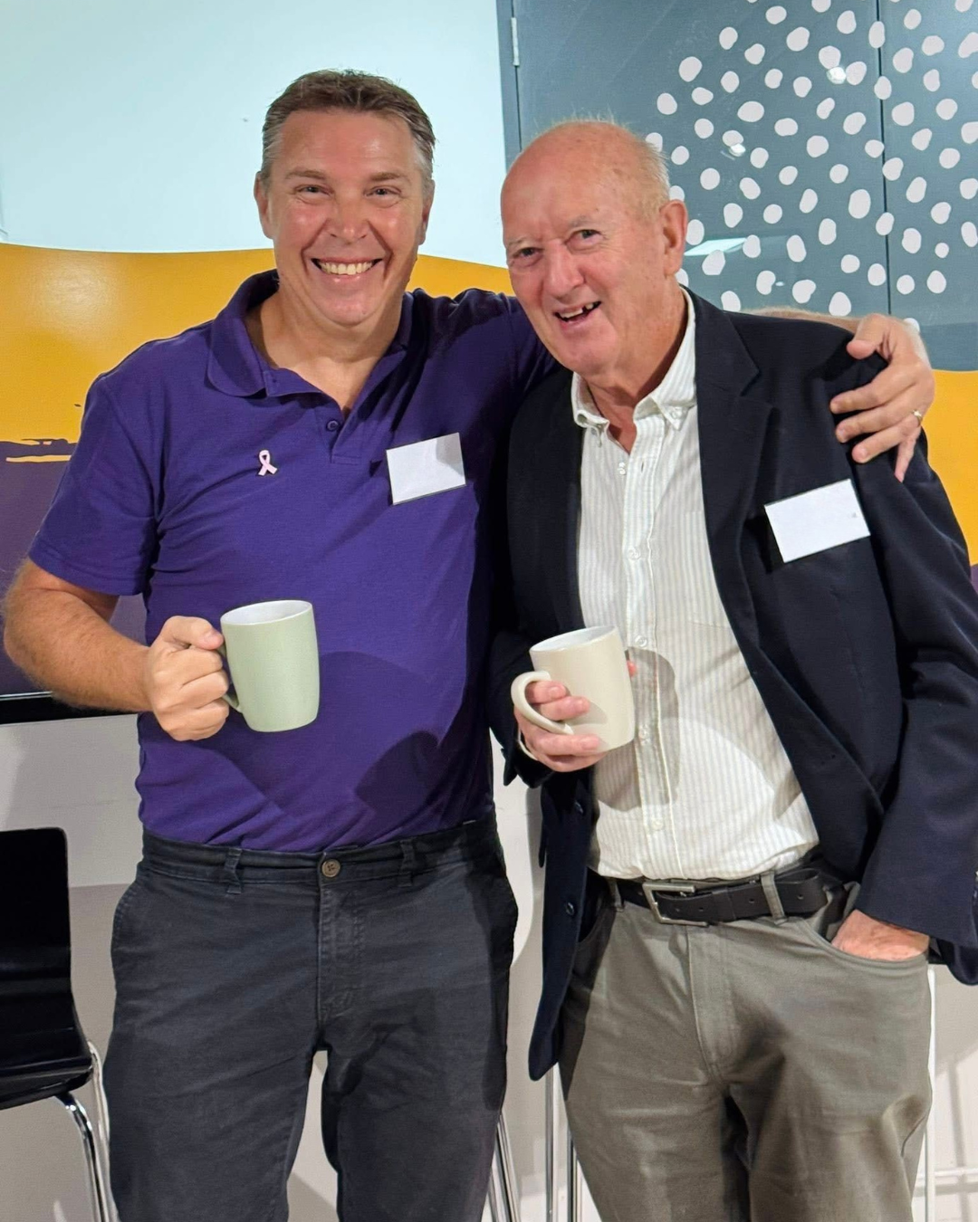 Two men standing together, smiling, and holding coffee mugs, at a fundraising event.