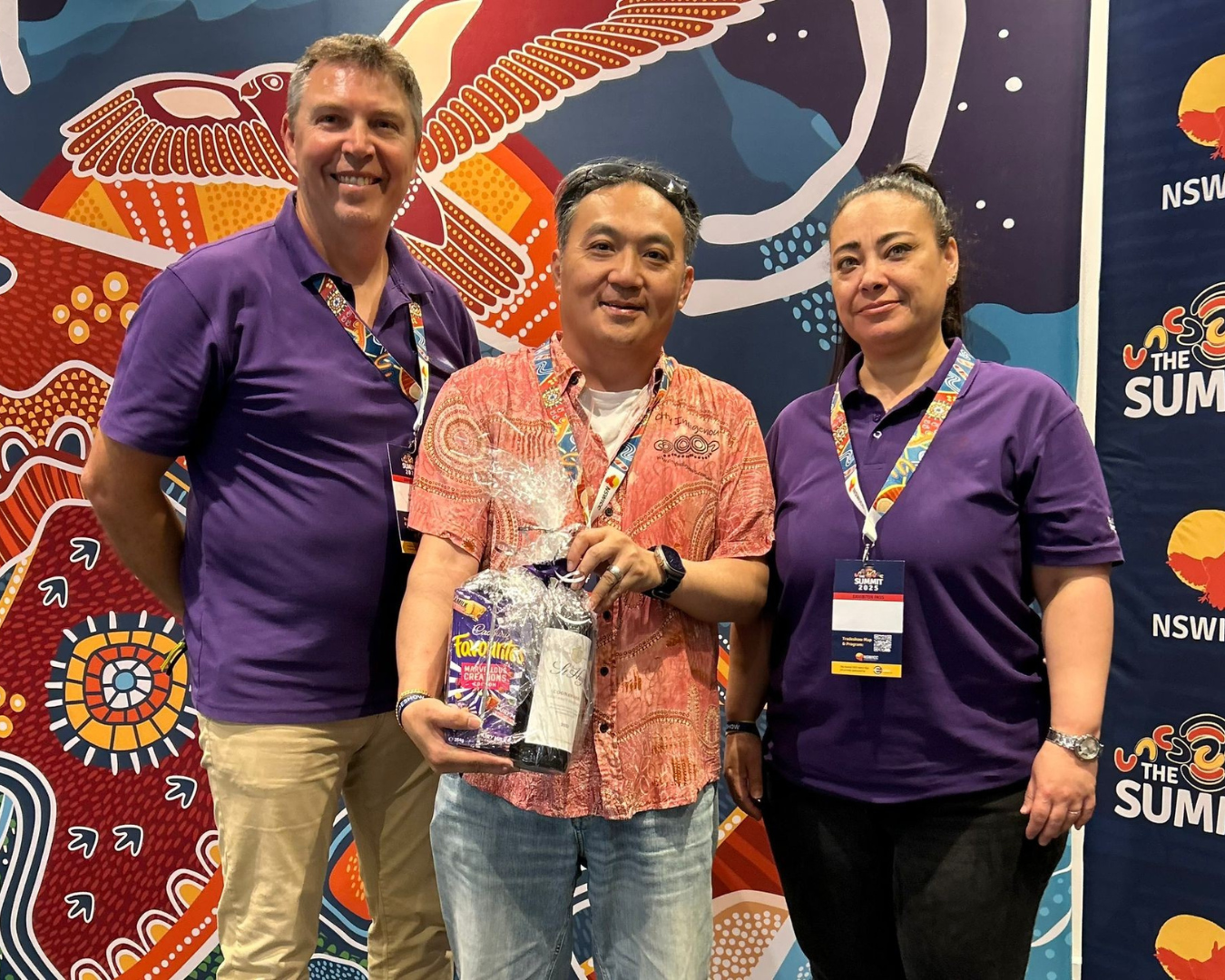 Three people standing in front of a colorful backdrop with Aboriginal Australian art designs, at a fundraising conference or event.