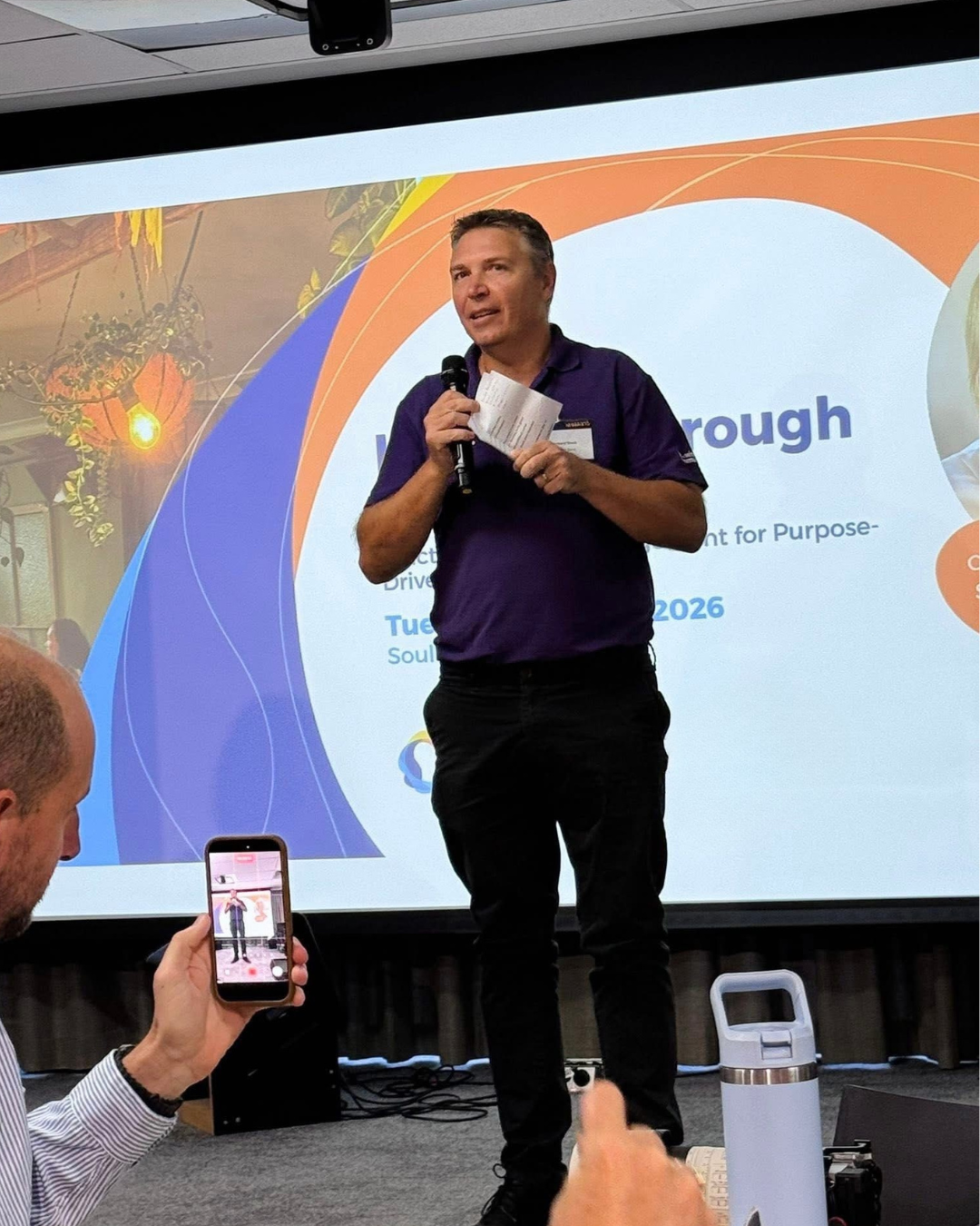 Man in purple shirt holding microphone and notes, speaking at a presentation, for a fundraising conference or event.