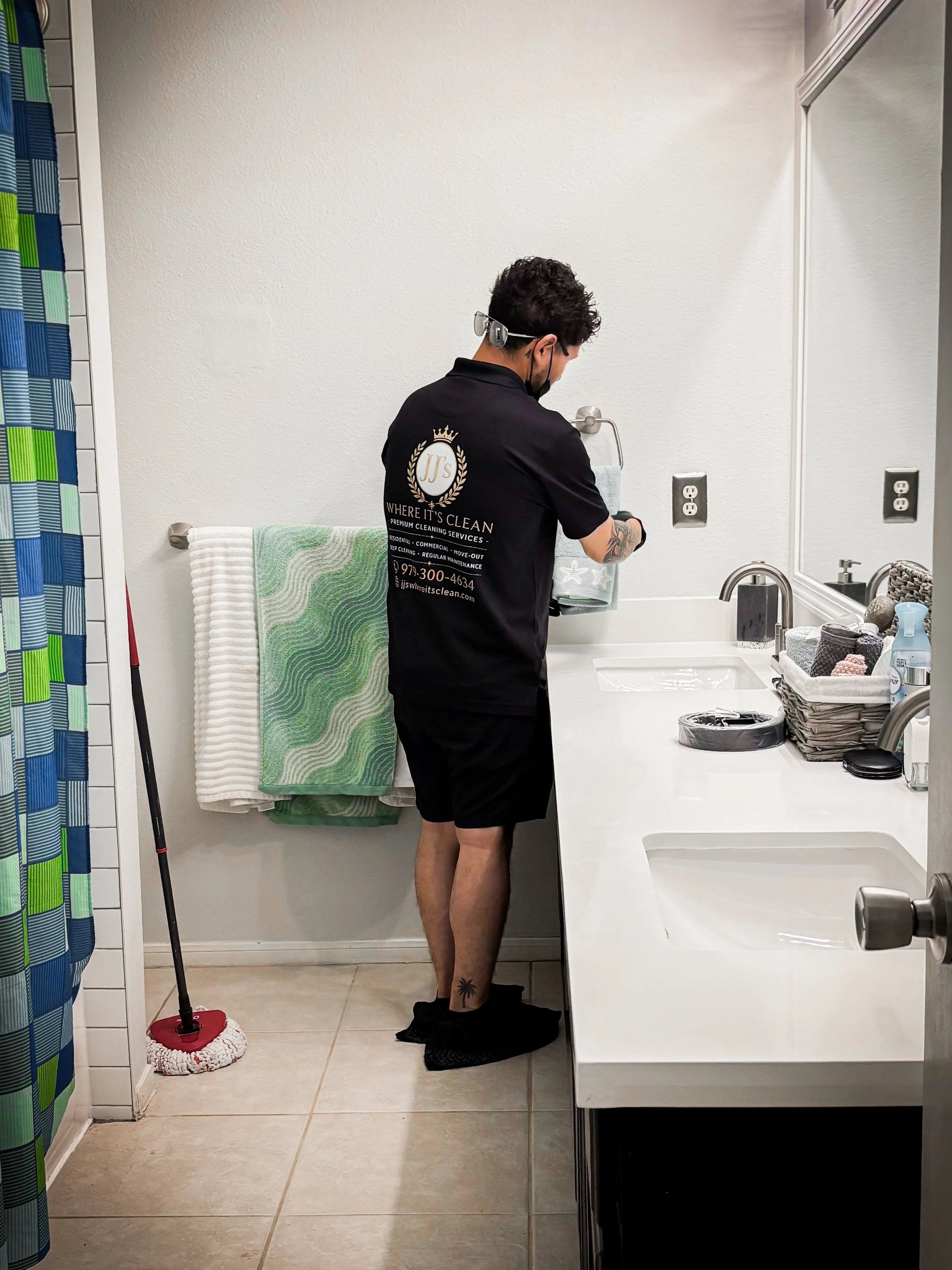 A man in a black cleaning service uniform handling a cloth in a bathroom, with a towel rack, sink, and bathroom accessories visible.