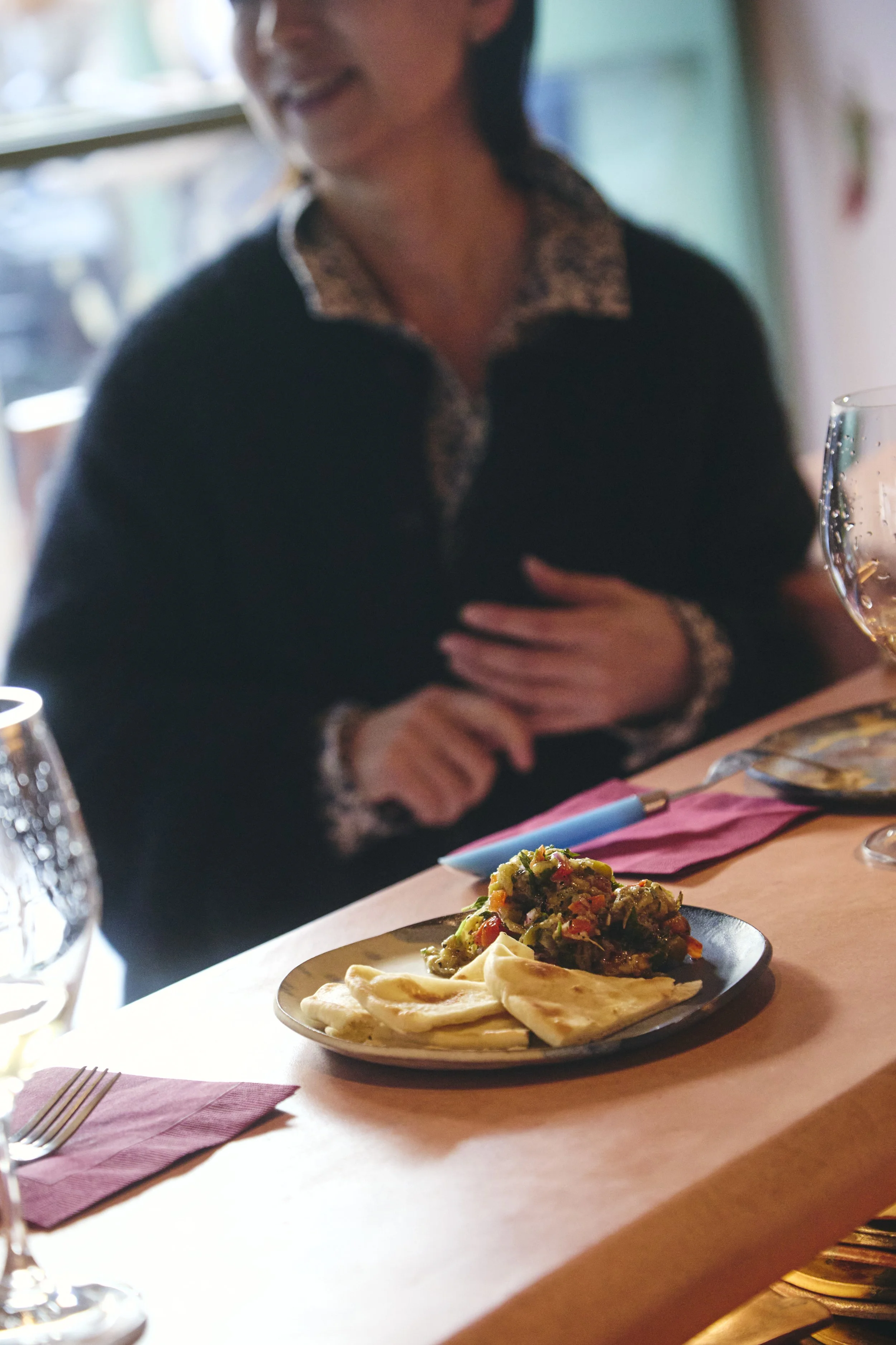 A woman smiling at a dining table with a plate of food, including pita bread and a vegetable dish, and a glass of wine, in a restaurant setting.