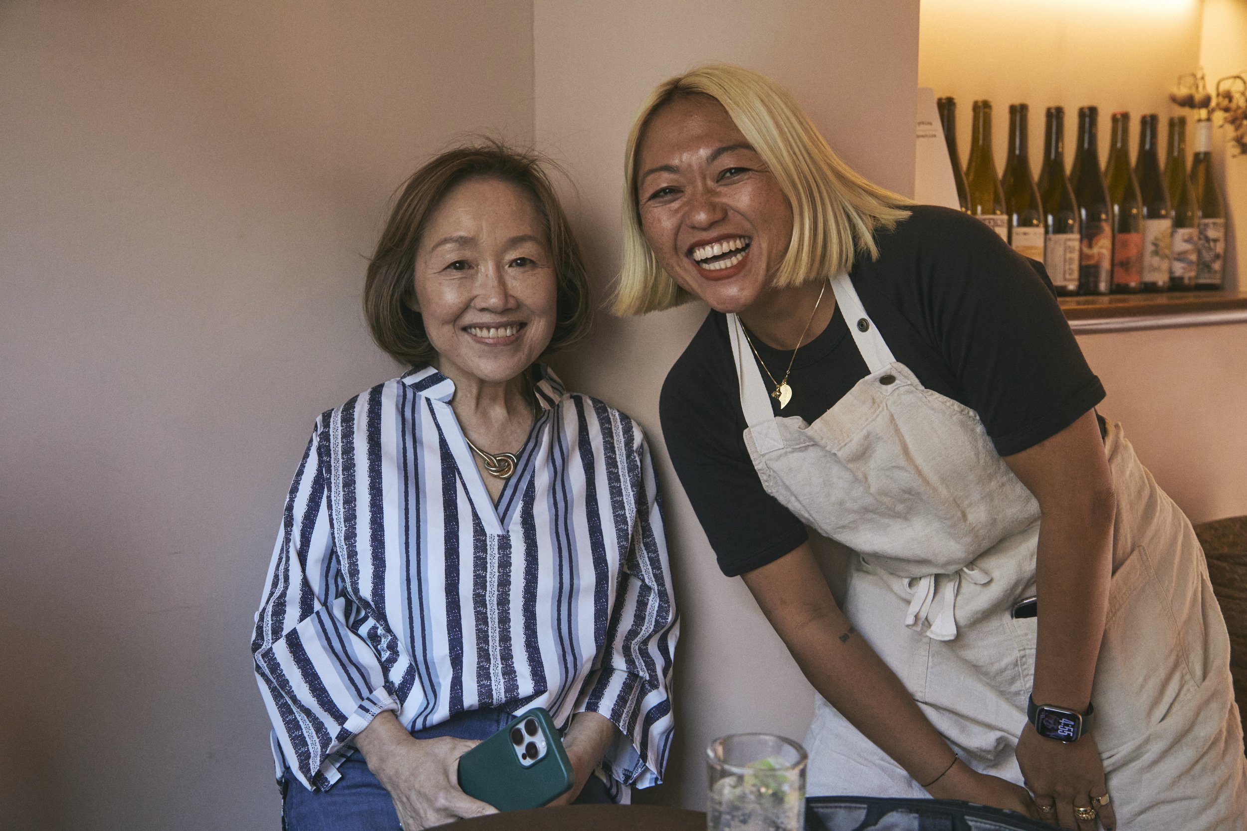 Two women smiling and posing together indoors. One is wearing a striped shirt and the other a black top with beige overalls. Bottles are on a shelf in the background.