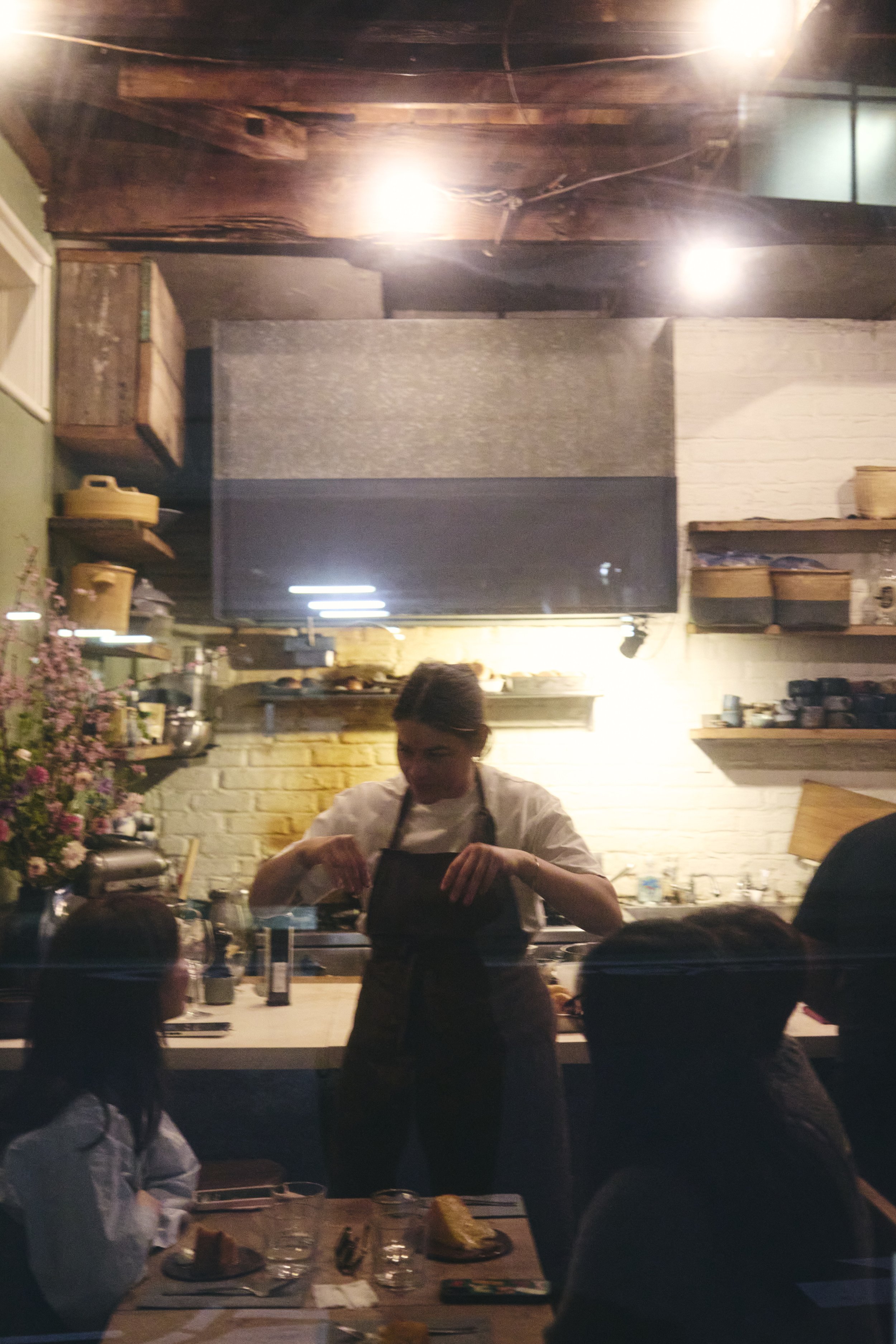 A woman is preparing food behind a counter in a restaurant. Several children are sitting at the counter with plates of food, listening and watching her. The restaurant has a cozy and rustic decor with wooden shelves, pots, and flowers, and a brick wa