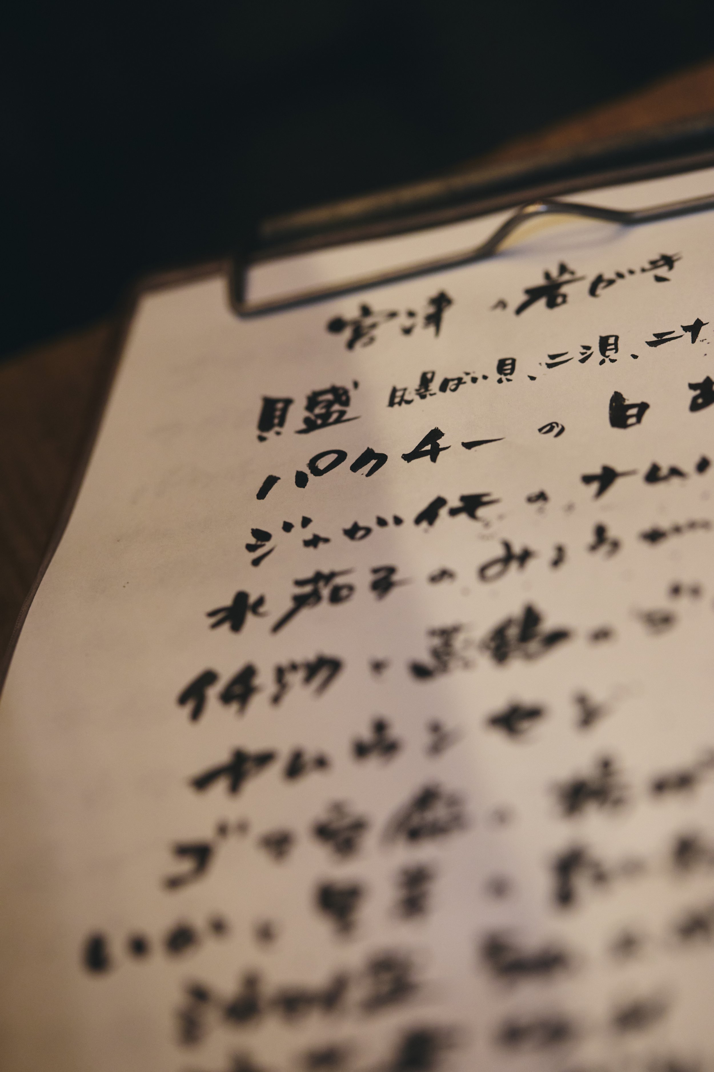 Close-up of a handwritten note in Japanese characters, with a black paperclip at the top left corner, slightly out of focus.