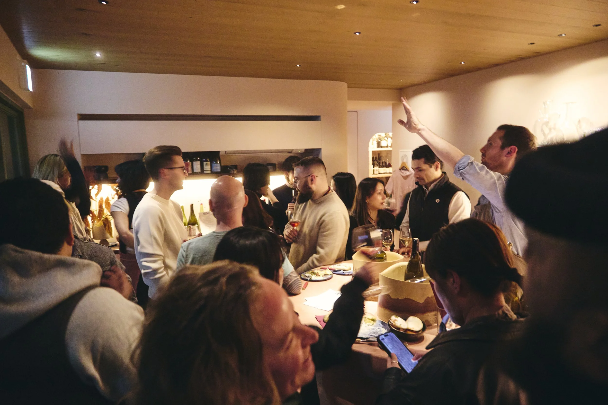 A crowded indoor gathering with people socializing and drinking, with a warm, cozy ambiance and dim lighting.