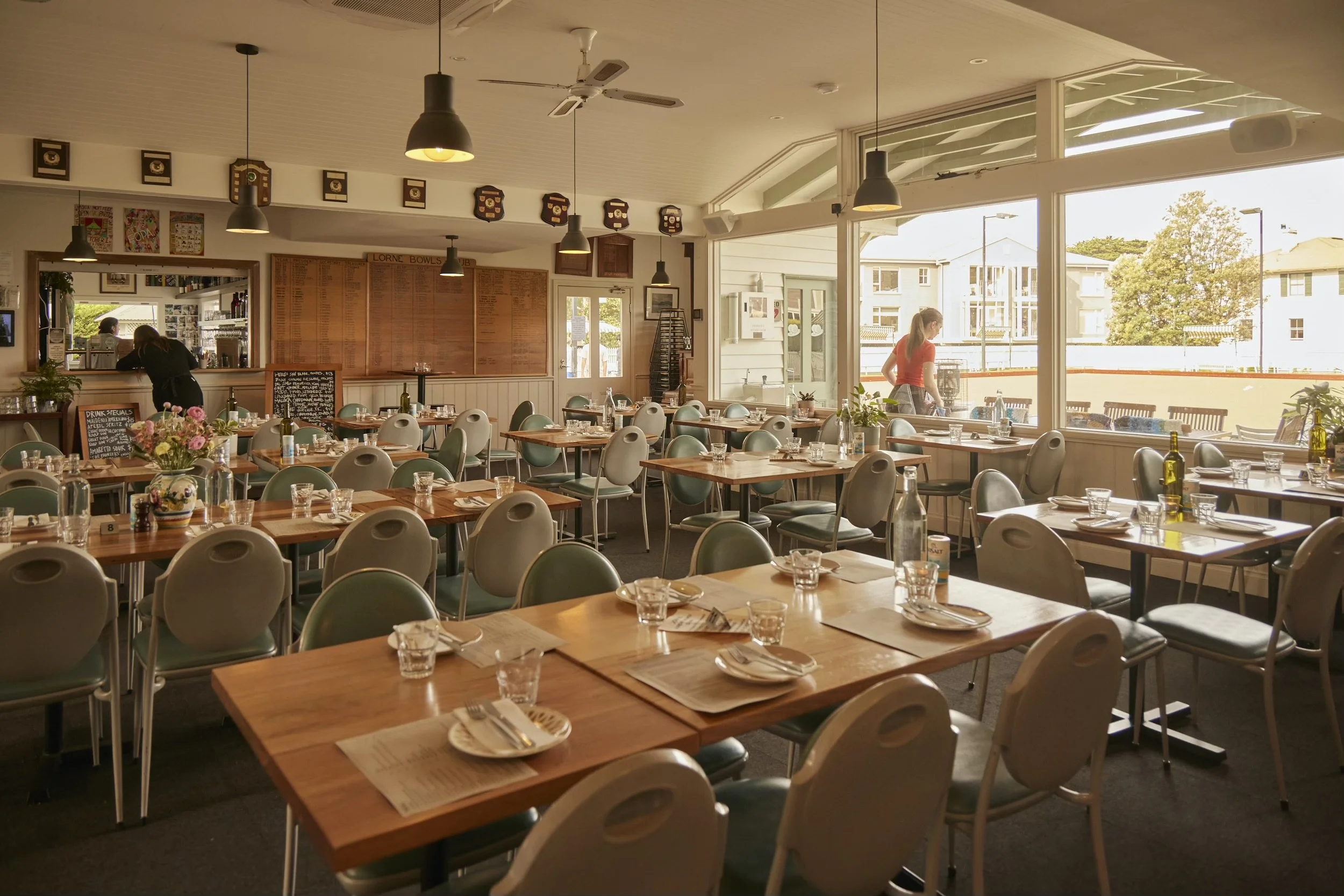 Interior of a restaurant with wooden tables set with glasses and utensils, large windows letting in natural light, and a few people, including waitstaff, inside.