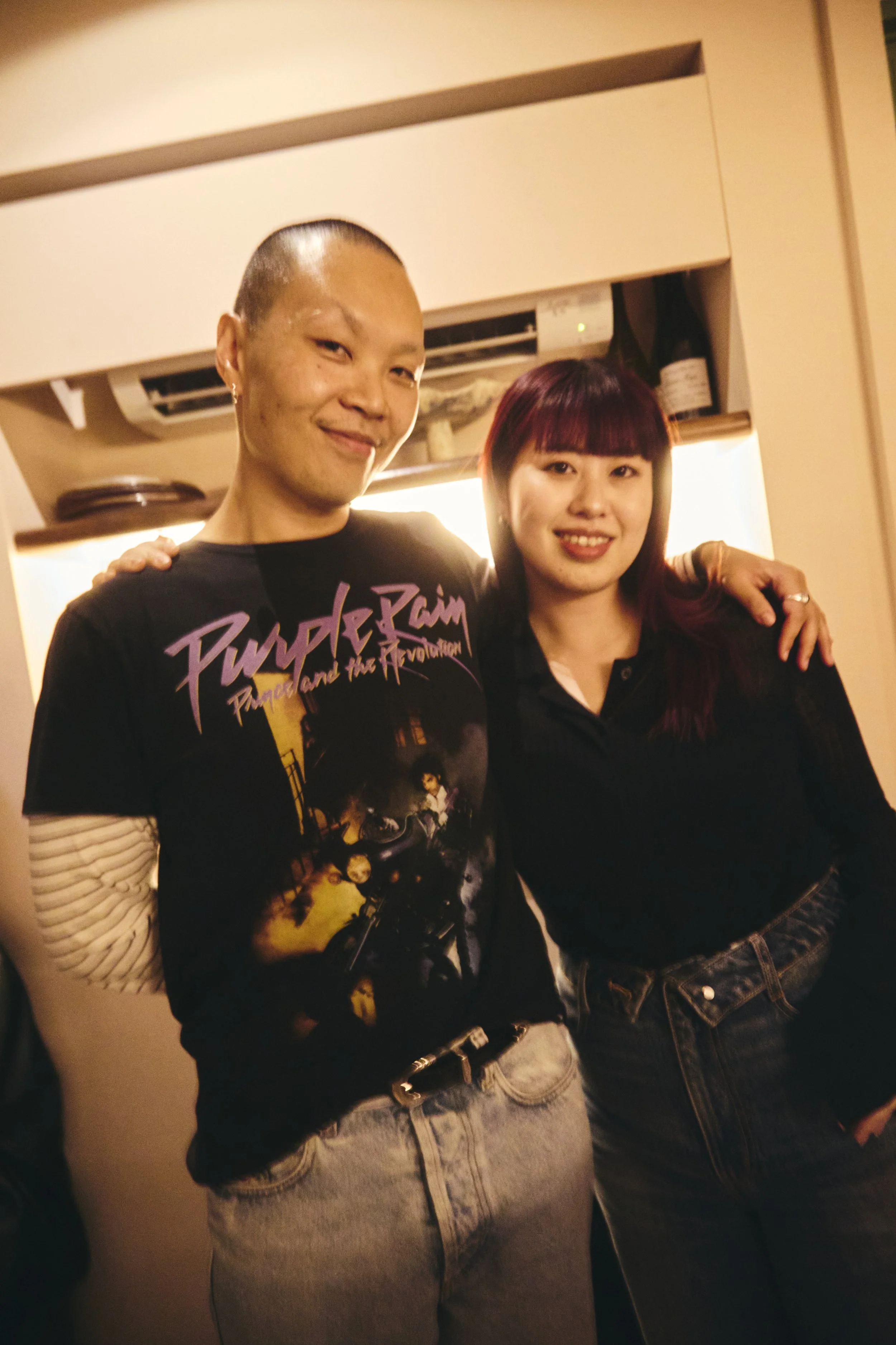 Two women posing together indoors in front of a beige wall and air conditioning unit. The woman on the left has short hair, earrings, a black graphic T-shirt, and light-washed jeans. The woman on the right has shoulder-length, reddish-purple hair, an