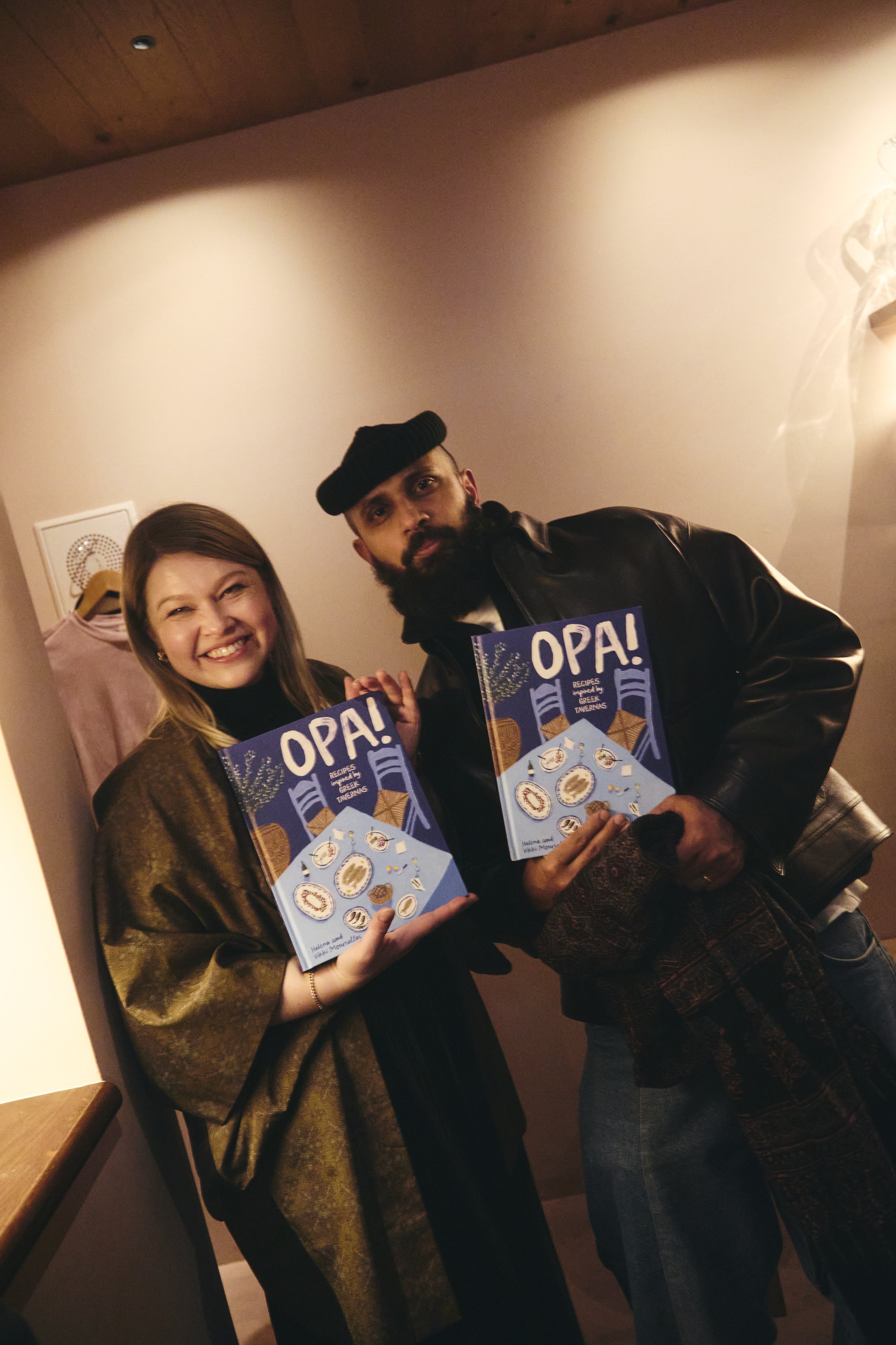 A smiling woman and a serious man holding a cookbook titled 'OPA! Recipes inspired by Greek tavernas' at a social gathering indoors. The woman has long brown hair and is wearing a brown shawl, while the man has a beard, mustache, and is wearing a bla