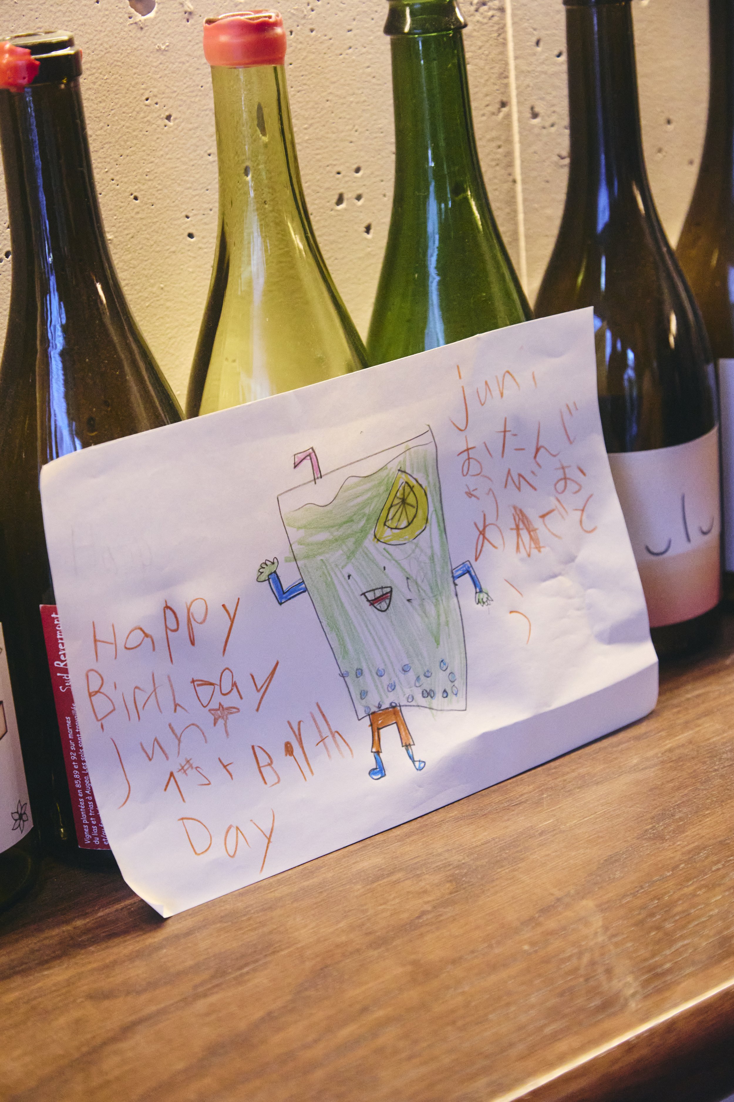 Colorful bottles of soda on a wooden surface with a handwritten birthday card featuring a drawing of a happy drink with a straw, lemon slice, and arms and legs.