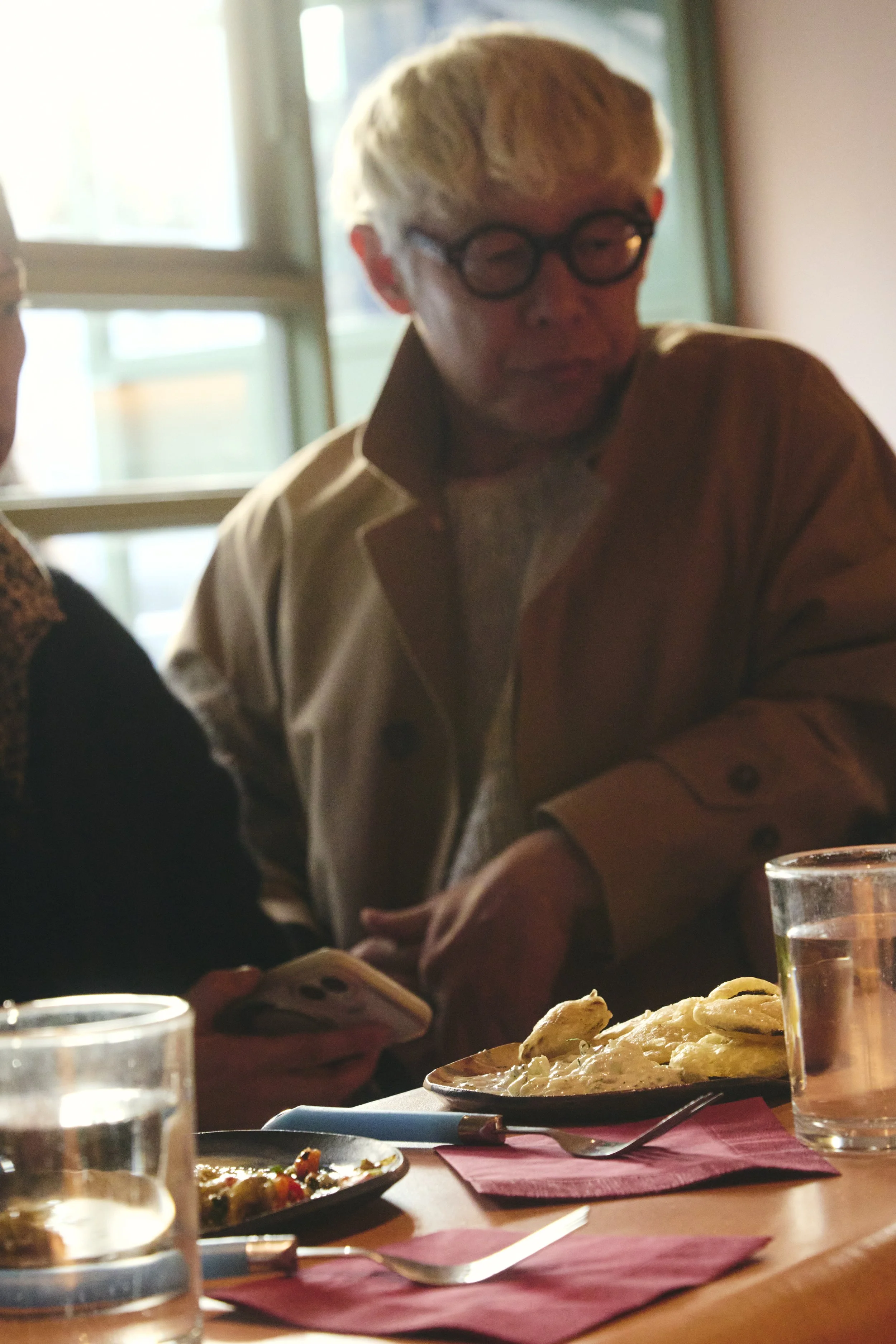 A person with white hair and glasses sitting at a table with plates of food, a glass of water, and pink napkins in a cozy, well-lit restaurant.