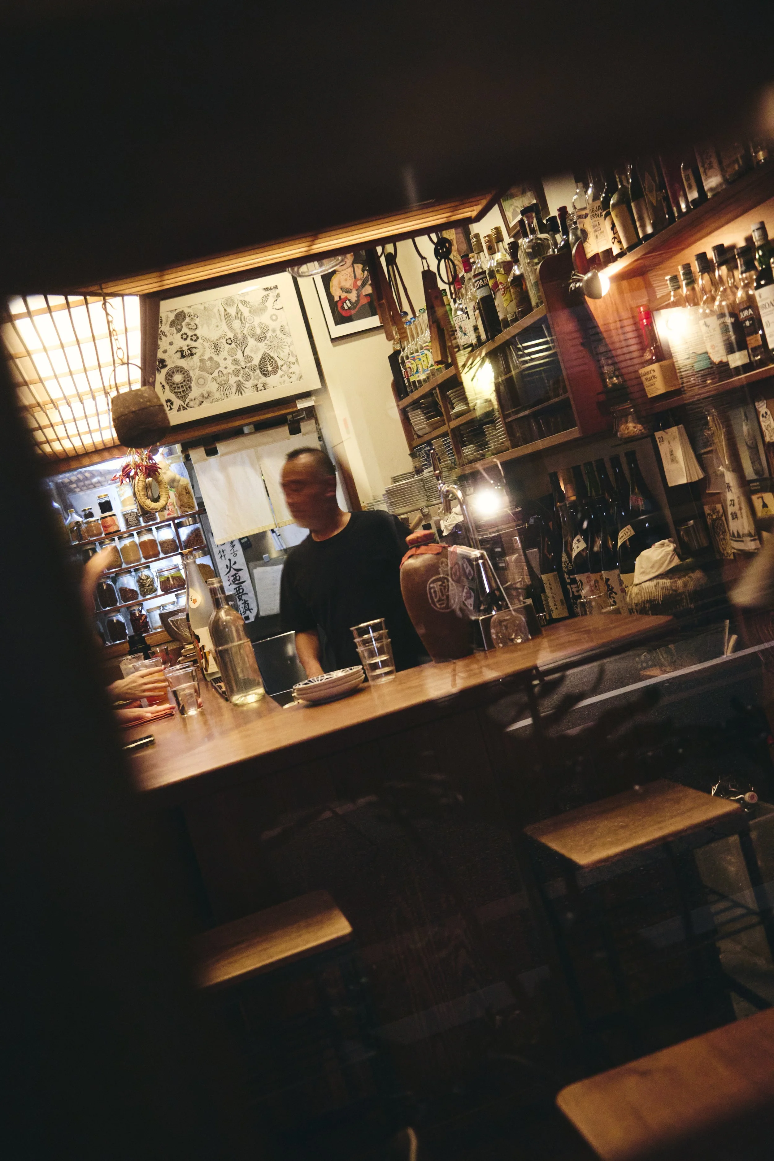 A bar counter with bottles of alcohol, glasses, and a bartender preparing drinks. Decor includes framed artwork and traditional Japanese decorations.