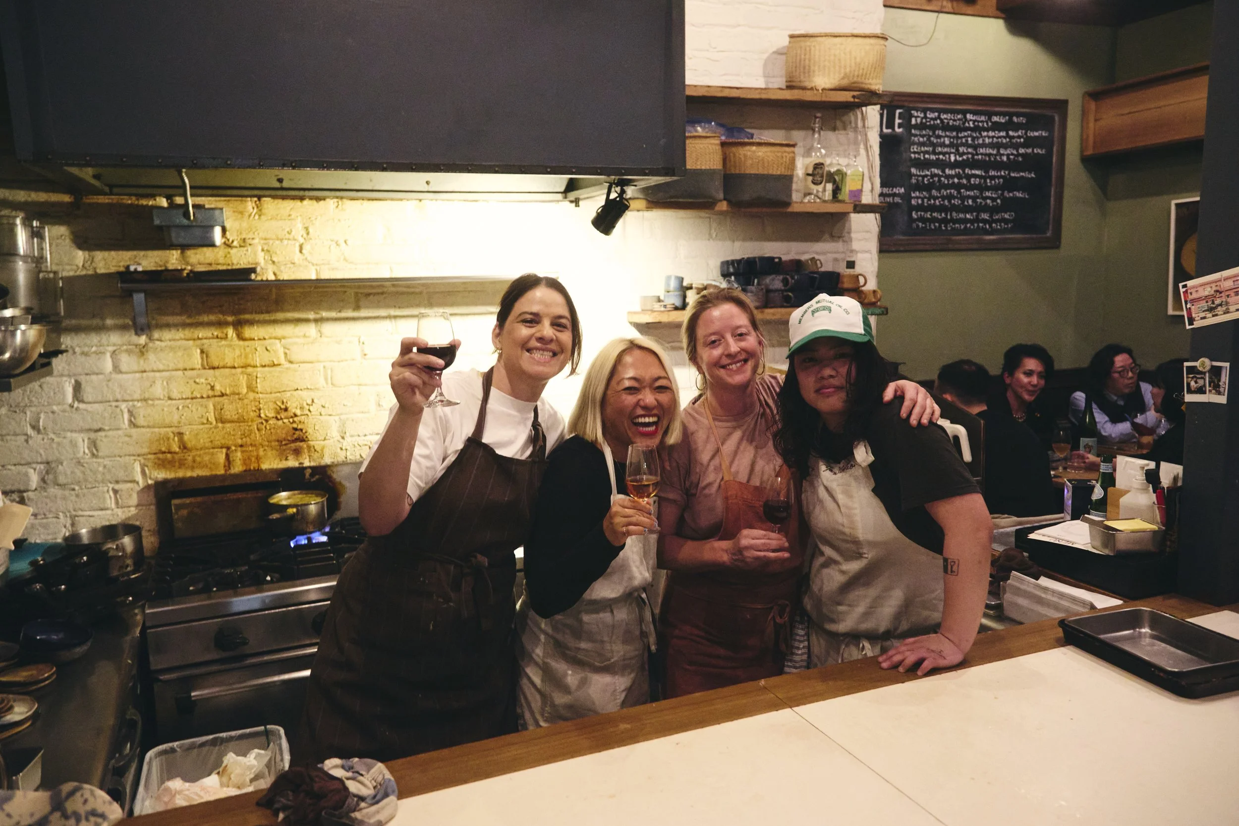 Four women standing together in a restaurant kitchen, smiling and holding wine glasses, with others dining in the background.