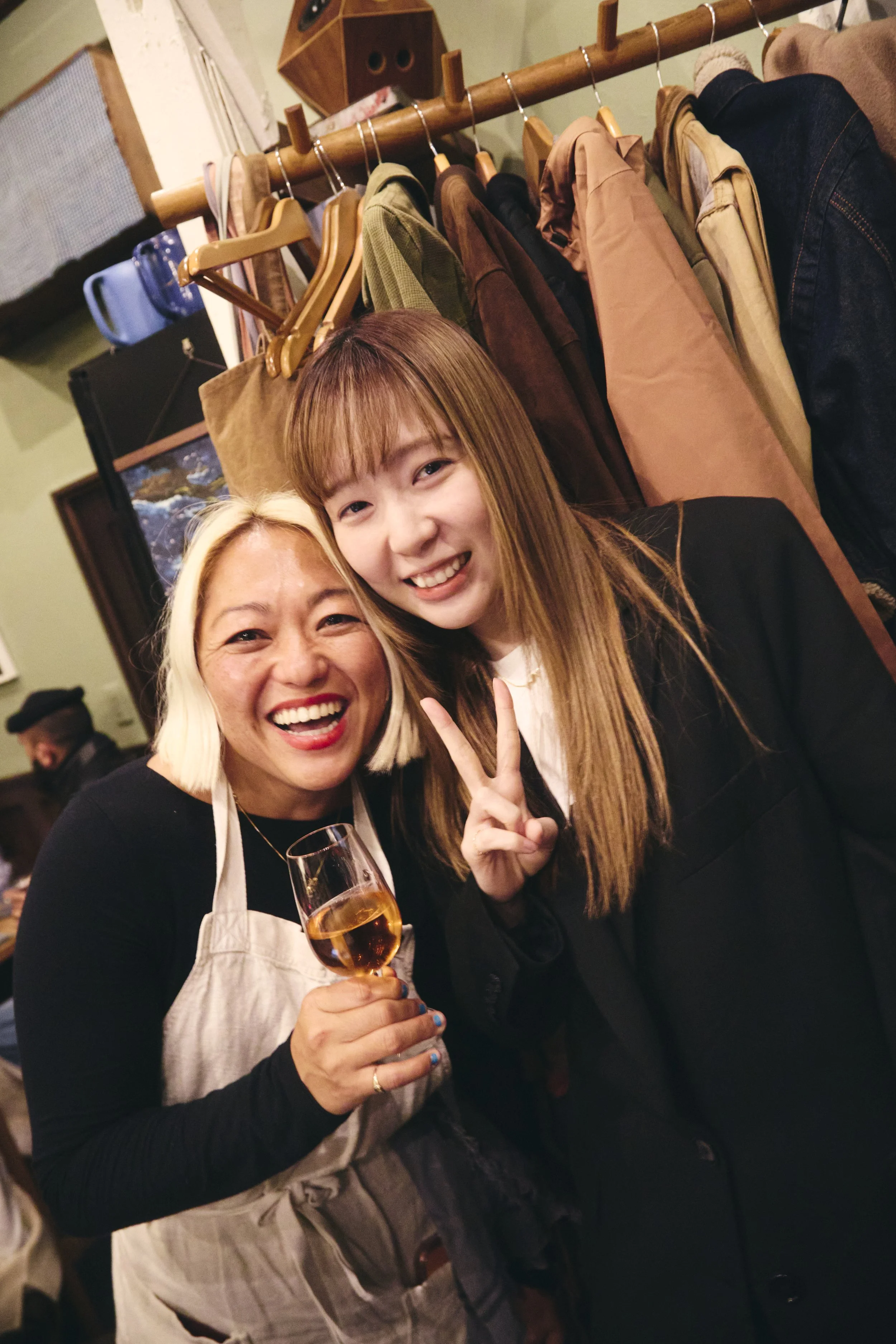 Two women smiling, one holding a glass of rosé wine, in a cozy indoor setting with a clothing rack, hanging jackets, and a decorative wooden shelf in the background.